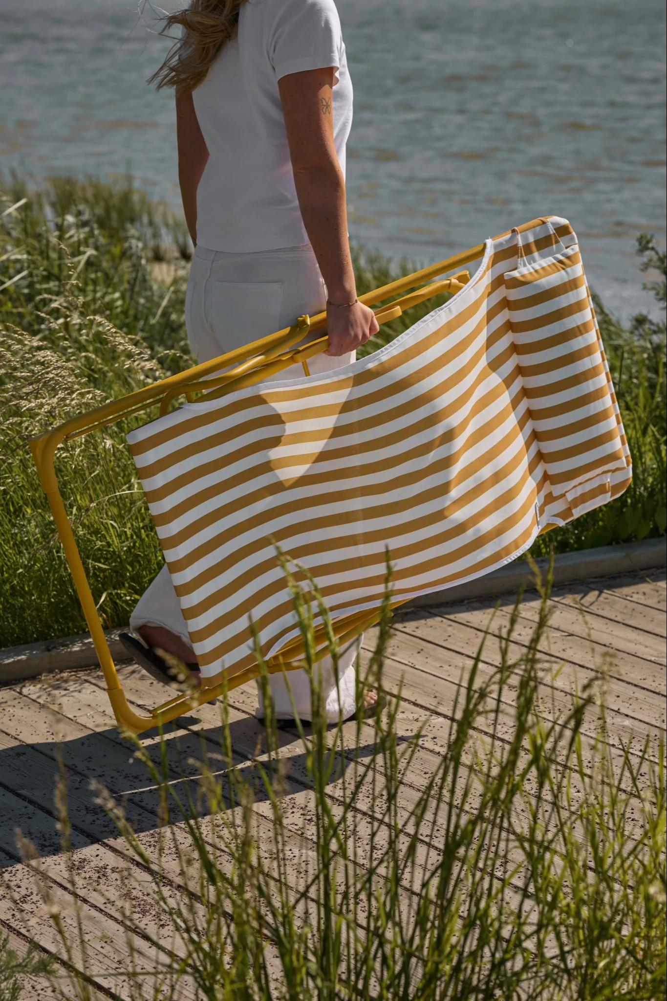 Persona de blanco lleva una silla de playa plegable a rayas amarillas y blancas en un paseo de madera con hierba alta junto al agua.