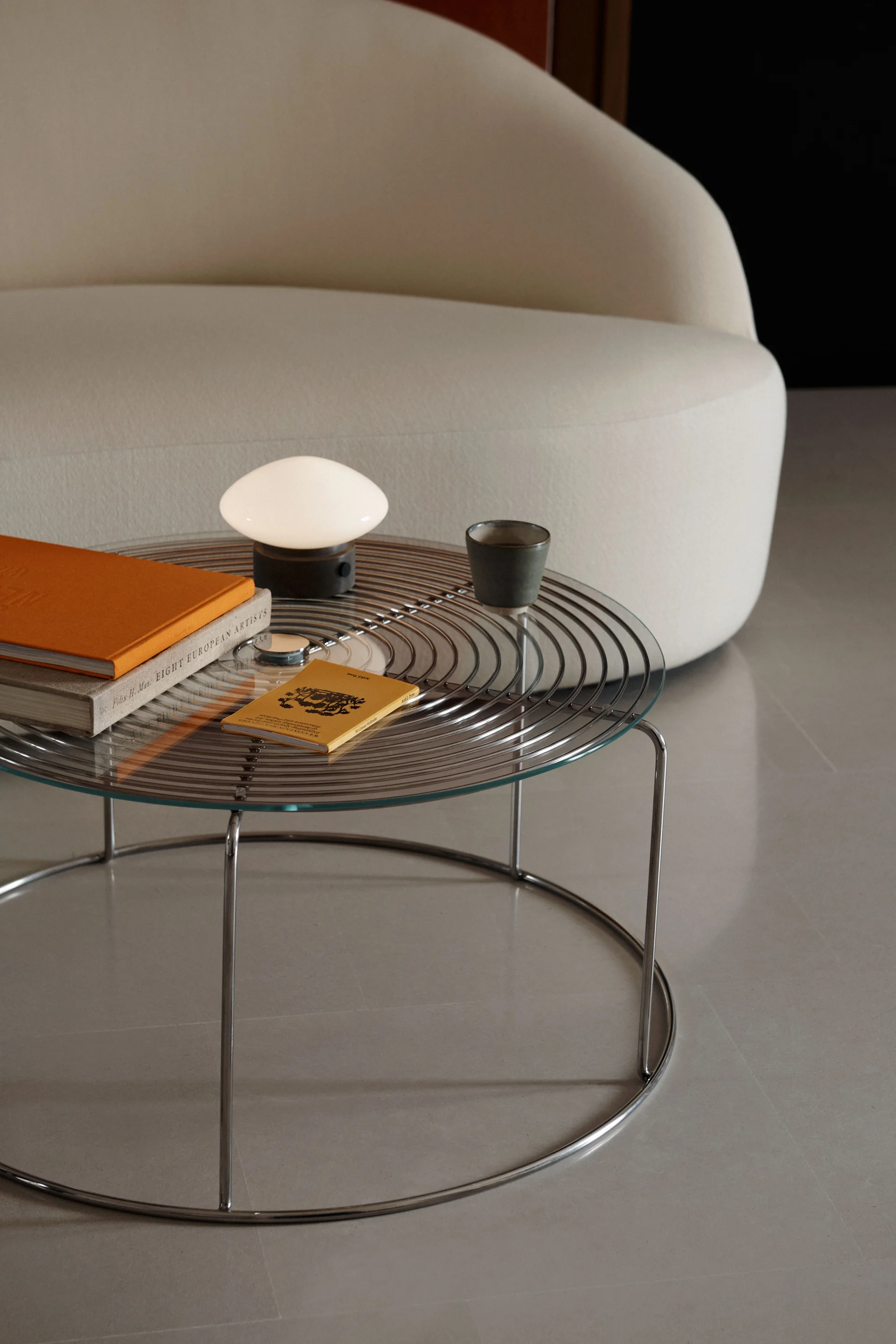 Una mesa de centro con tapa de cristal y base cromada sostiene una lámpara blanca con forma de hongo, un libro naranja y una taza oscura junto a un sofá curvo color crema.