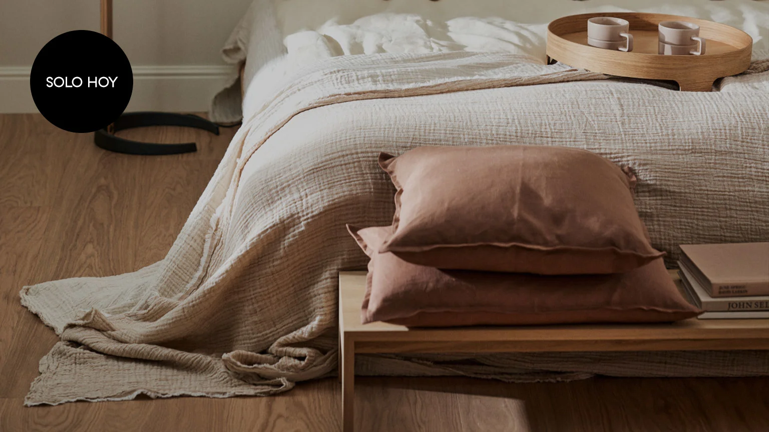 Cama con edredón beige claro, cojines marrones en una mesa de noche de madera, y una bandeja con tazas sobre la cama. Etiqueta 'SOLO HOY'.