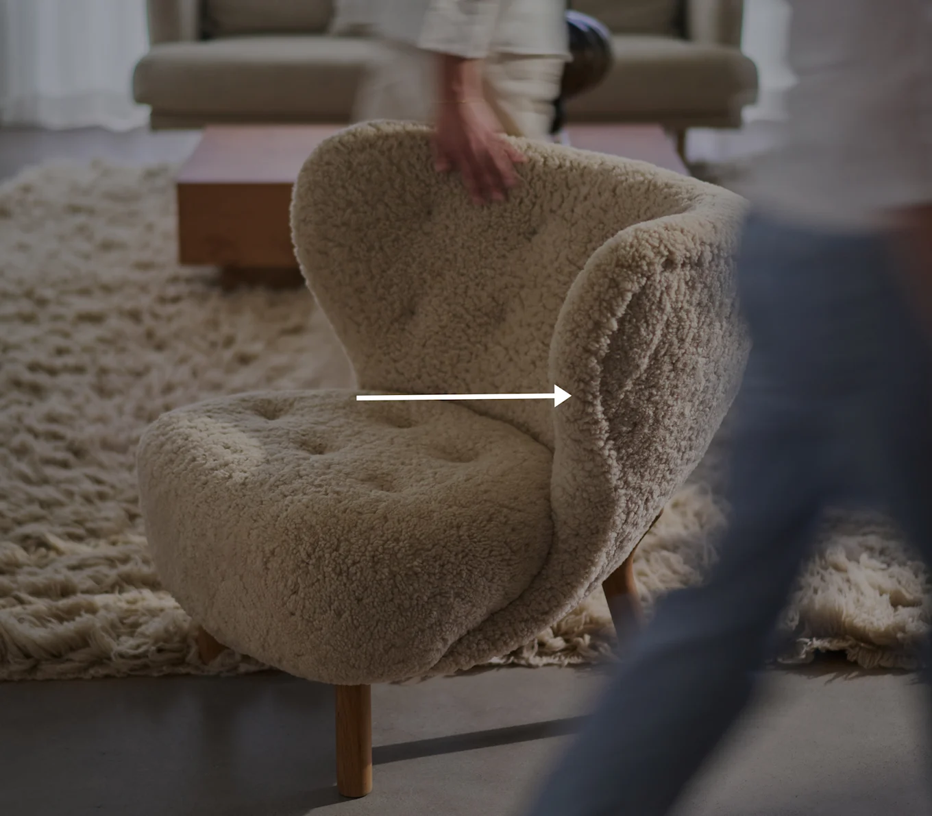 Sillón cremoso y mullido con patas de madera en una sala de estar, una mano borrosa toca el respaldo del sillón.