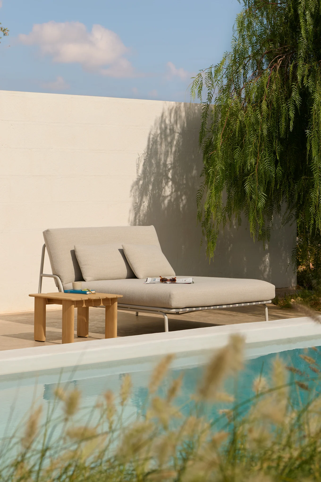 Tumbona gris claro con almohadas, una mesa auxiliar de madera y una revista junto a una piscina.