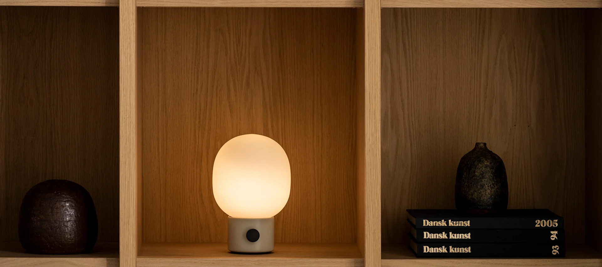 En una estantería descansa una luminosa lámpara de mesa JWDA de Audo Copenhagen.