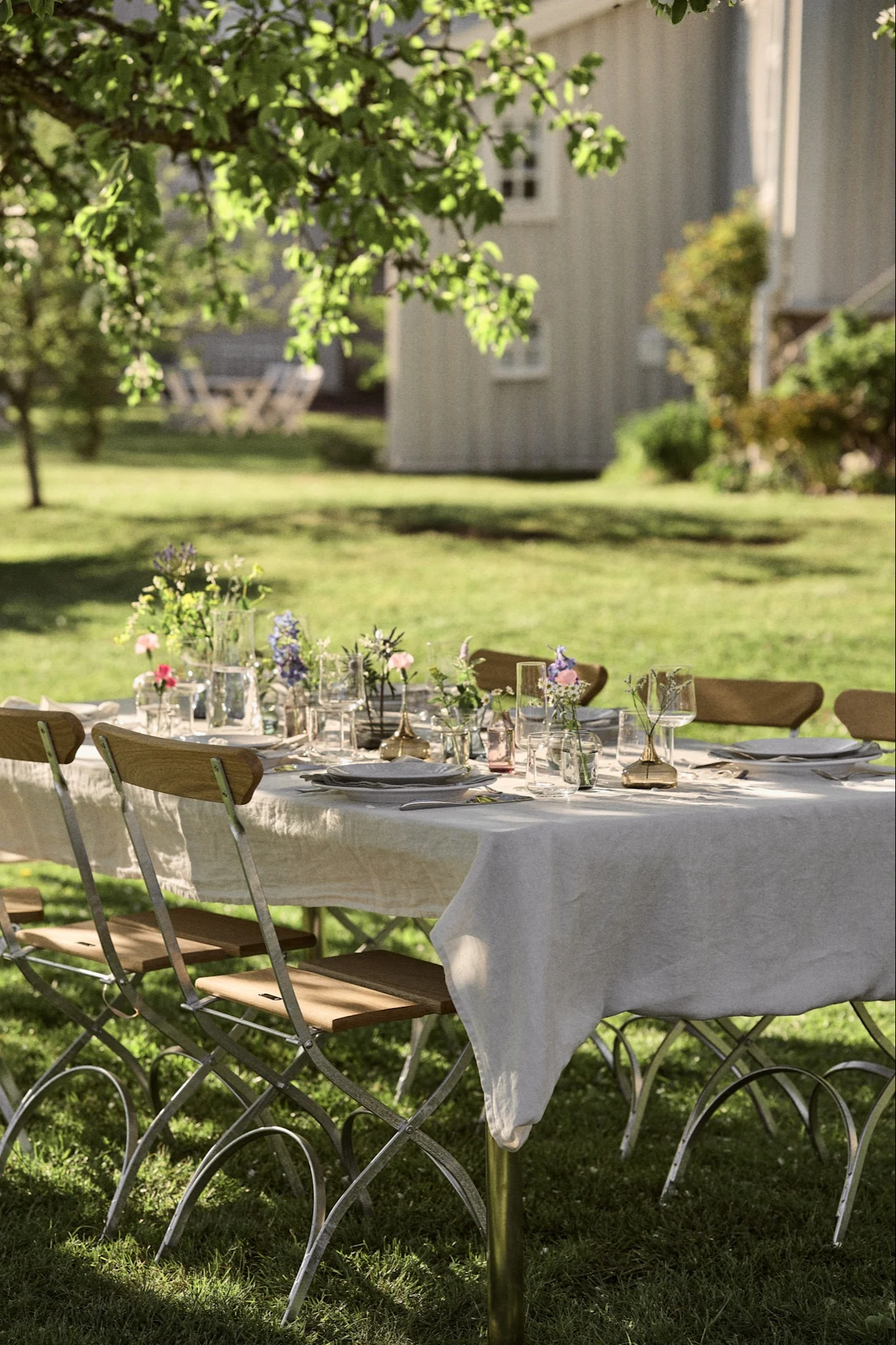 Una mesa de comedor al aire libre con platos blancos, vasos transparentes y pequeños ramos de flores sobre un mantel de lino en un jardín soleado.