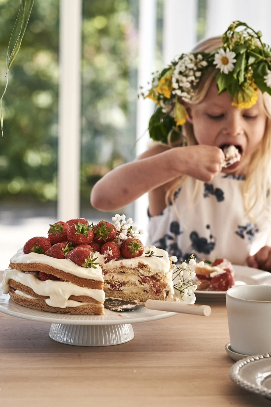 Prepara una mesa sencilla para el Midsummer y sirve la tarta de fresas en un plato blanco, como en la imagen, donde una niña con una corona de flores se come la tarta.