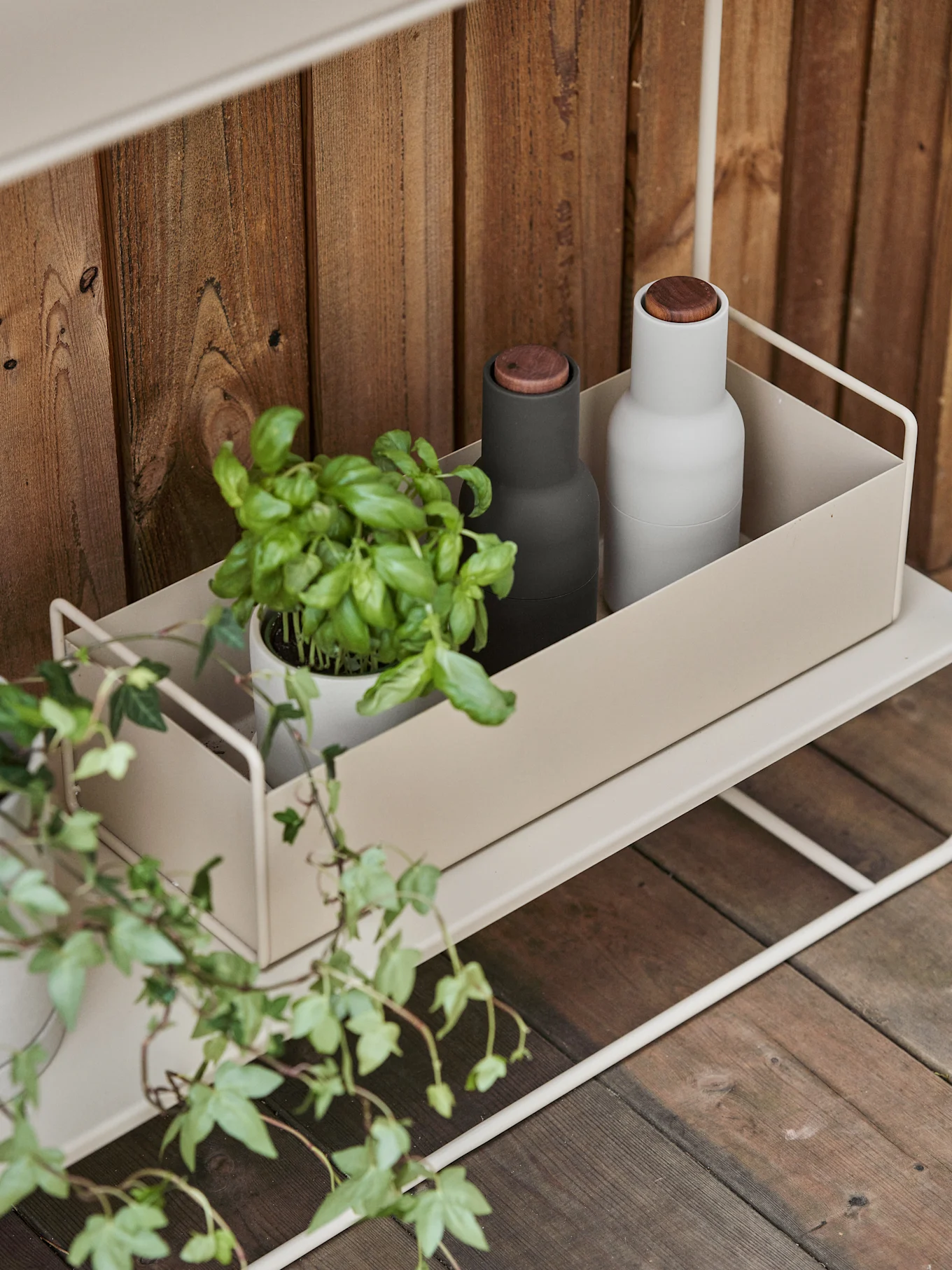Un estante de metal beige claro con un recipiente que contiene una planta de albahaca, un molinillo negro y un molinillo blanco sobre un fondo de madera.