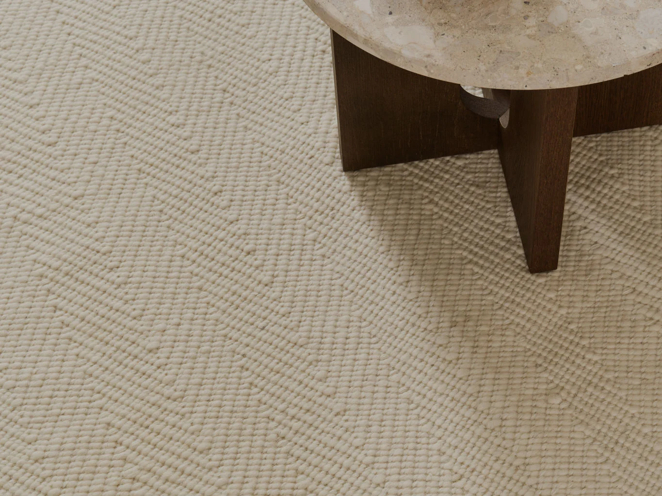 Primer plano de una alfombra texturizada color crema con patrón de chevrón, junto a una mesa con tablero de piedra.