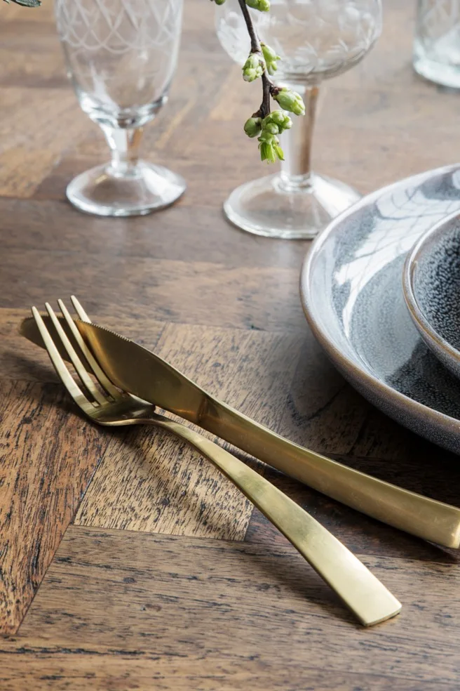 Tenedor y cuchillo dorados sobre una mesa de madera junto a platos de cerámica y vasos con dibujos, con una pequeña rama de brotes verdes.