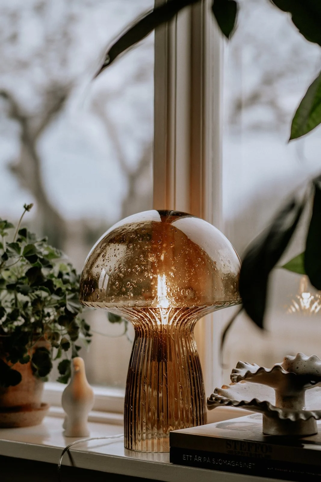 La lámpara de mesa Fungo marrón de Globen Lighting se coloca en el alféizar de una ventana rodeada de plantas: la decoración boho perfecta.