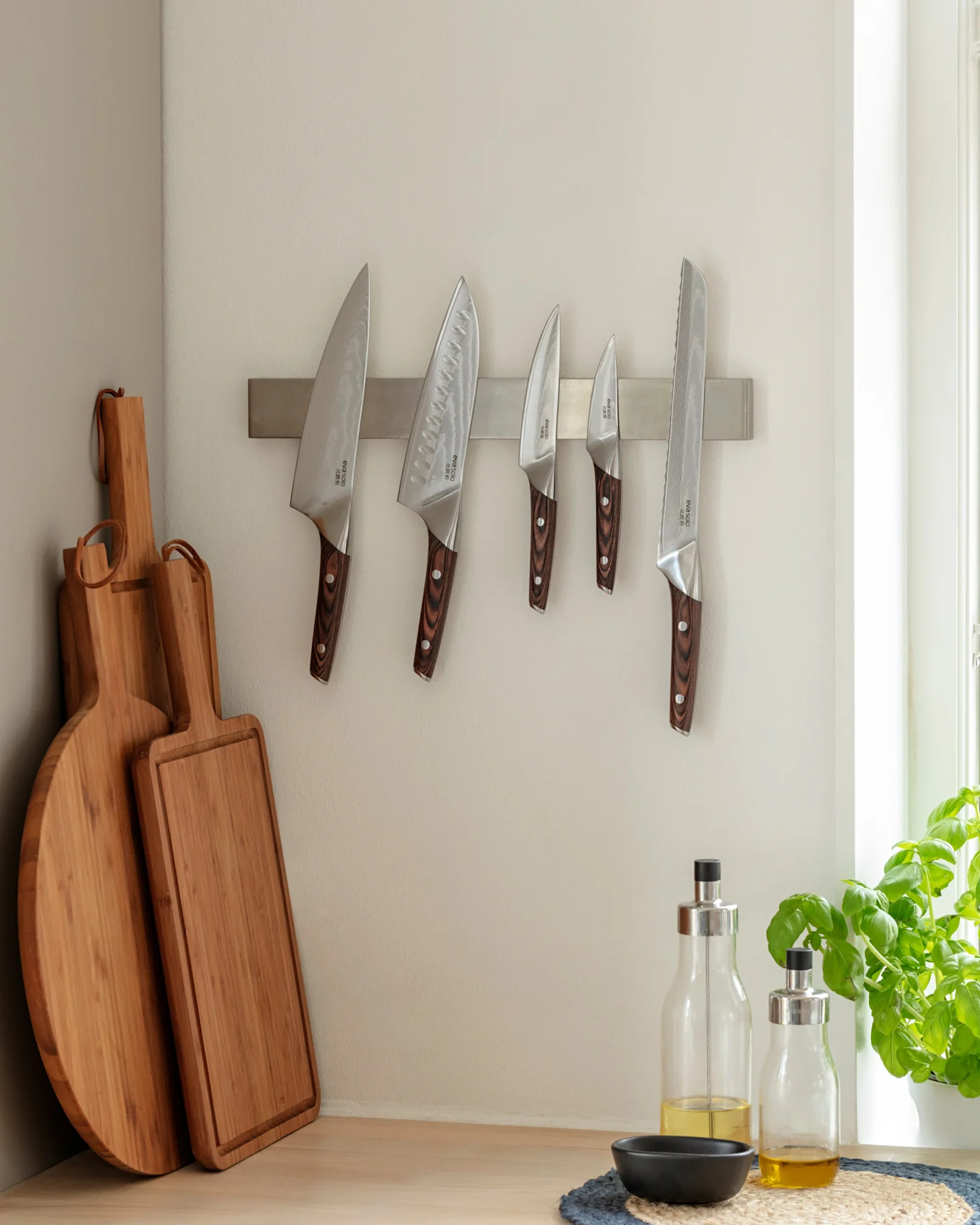 Rincón de cocina con cinco cuchillos en una tira magnética, dos tablas de cortar de madera y dos botellas de aceite de oliva en una encimera de madera junto a una planta de albahaca.