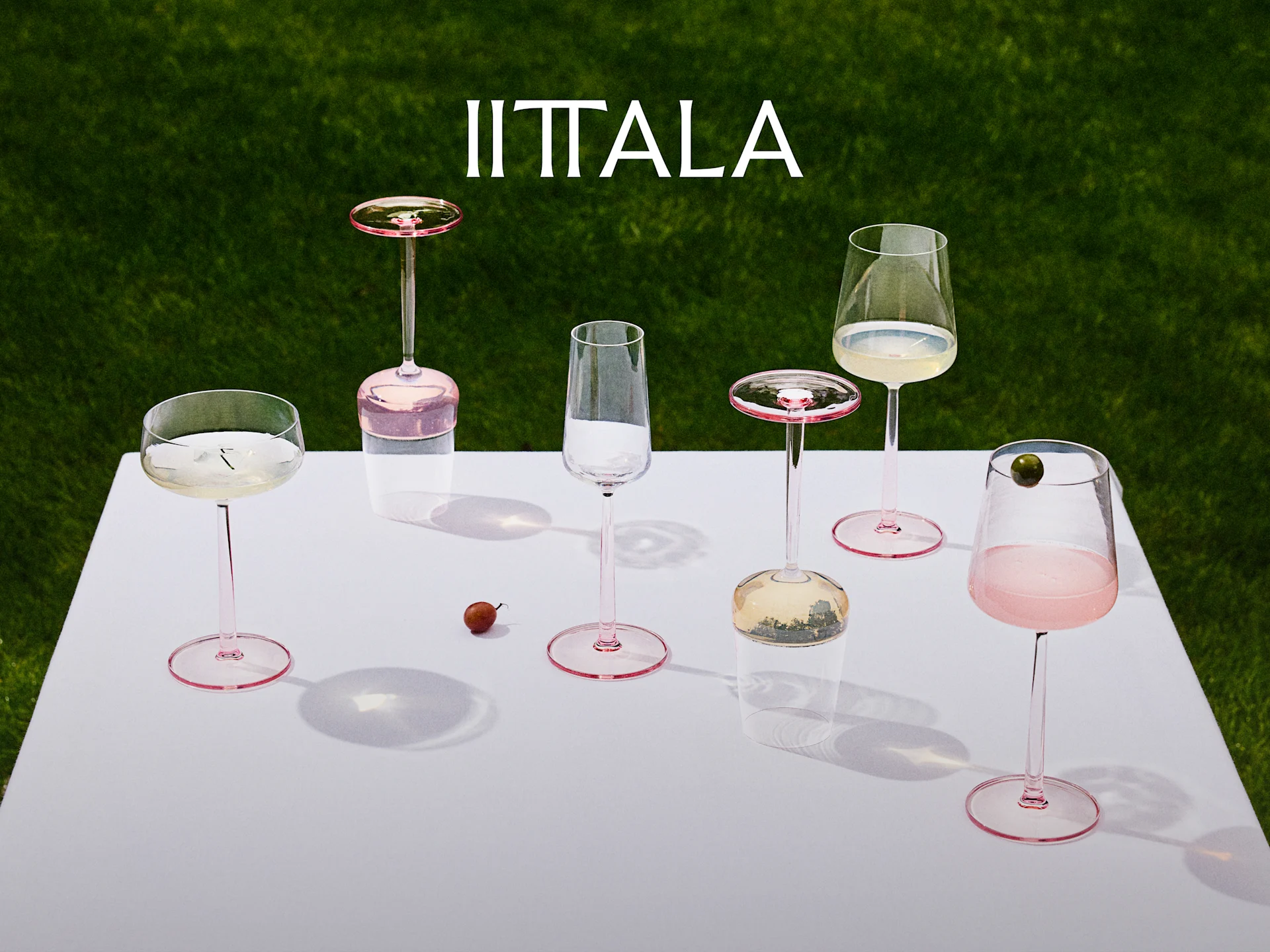 Logotipo de Iittala sobre una colección de copas con tallo rosa en una mesa blanca con fondo de césped verde, algunas con bebidas.