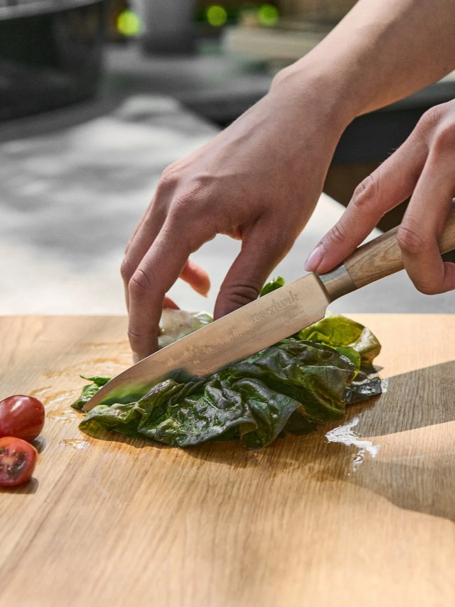 Manos cortando lechuga verde fresca en una tabla de madera con un cuchillo, junto a tomates cherry en rodajas.