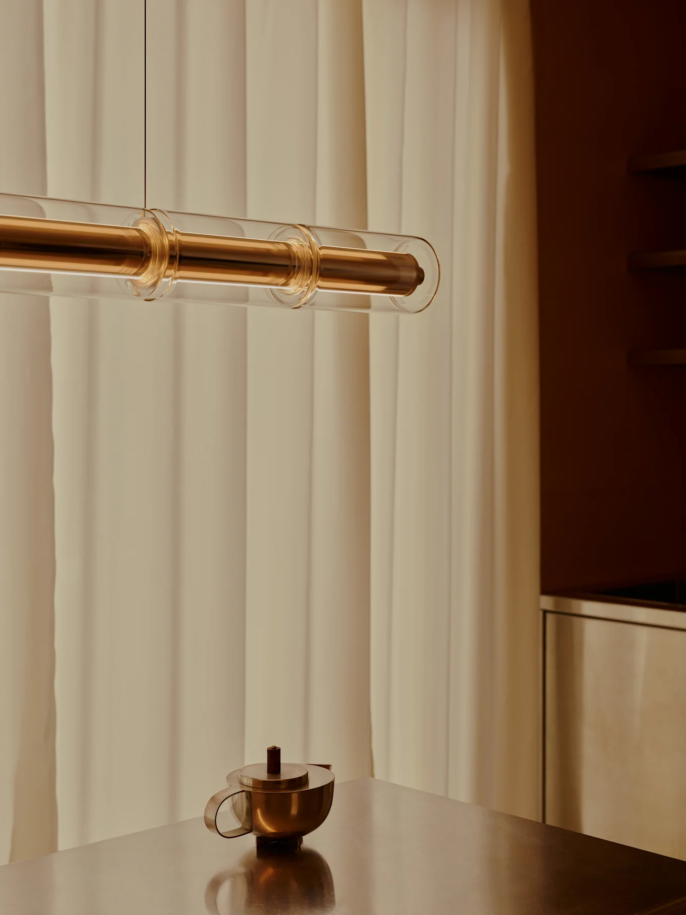 Una lámpara colgante moderna de latón y tubos de vidrio cuelga sobre una tetera de latón en una mesa metálica, con cortinas claras al fondo.