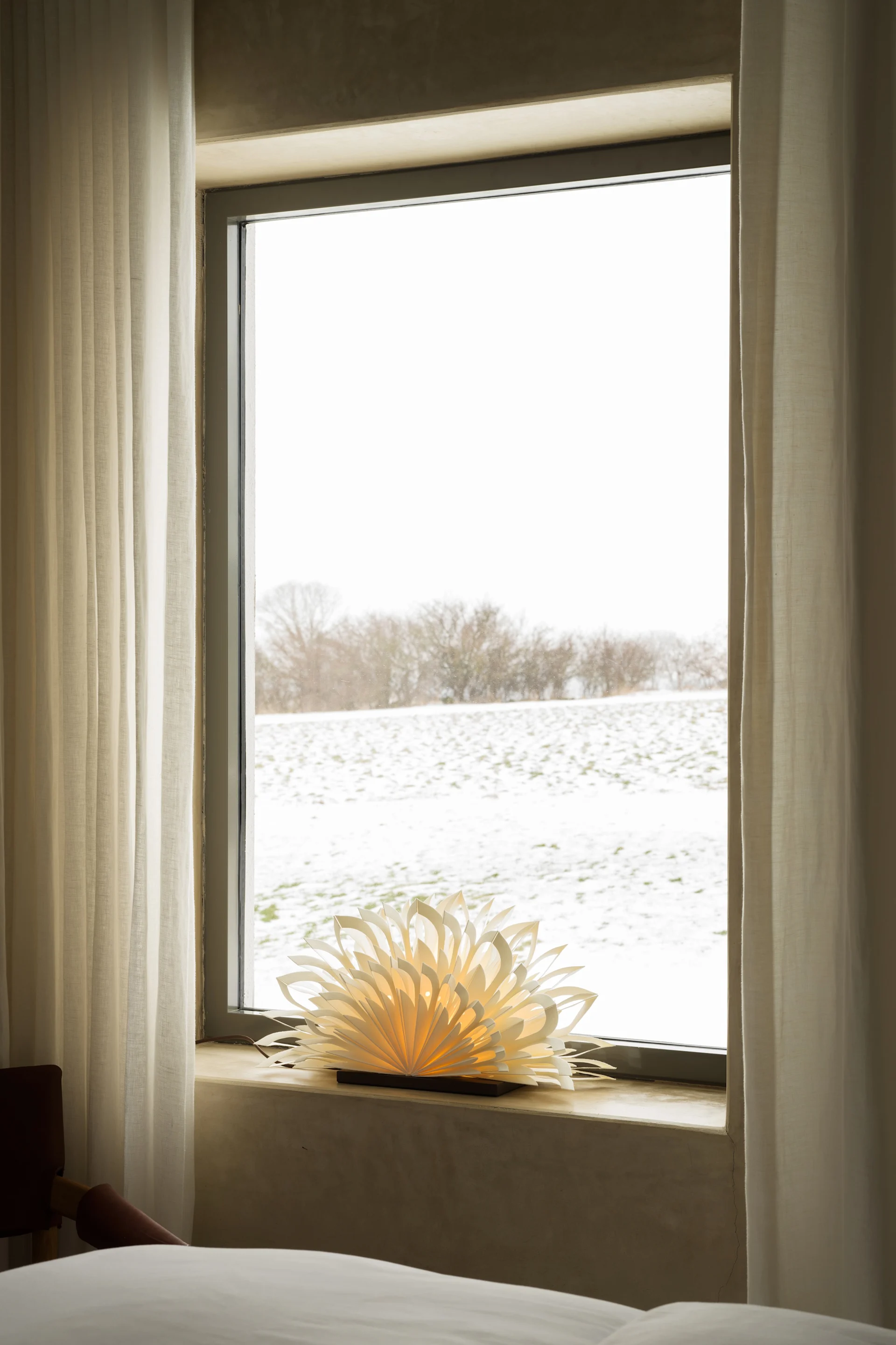 Ventana con vistas a un campo nevado, cortinas blancas y una lámpara de papel brillante en el alféizar.