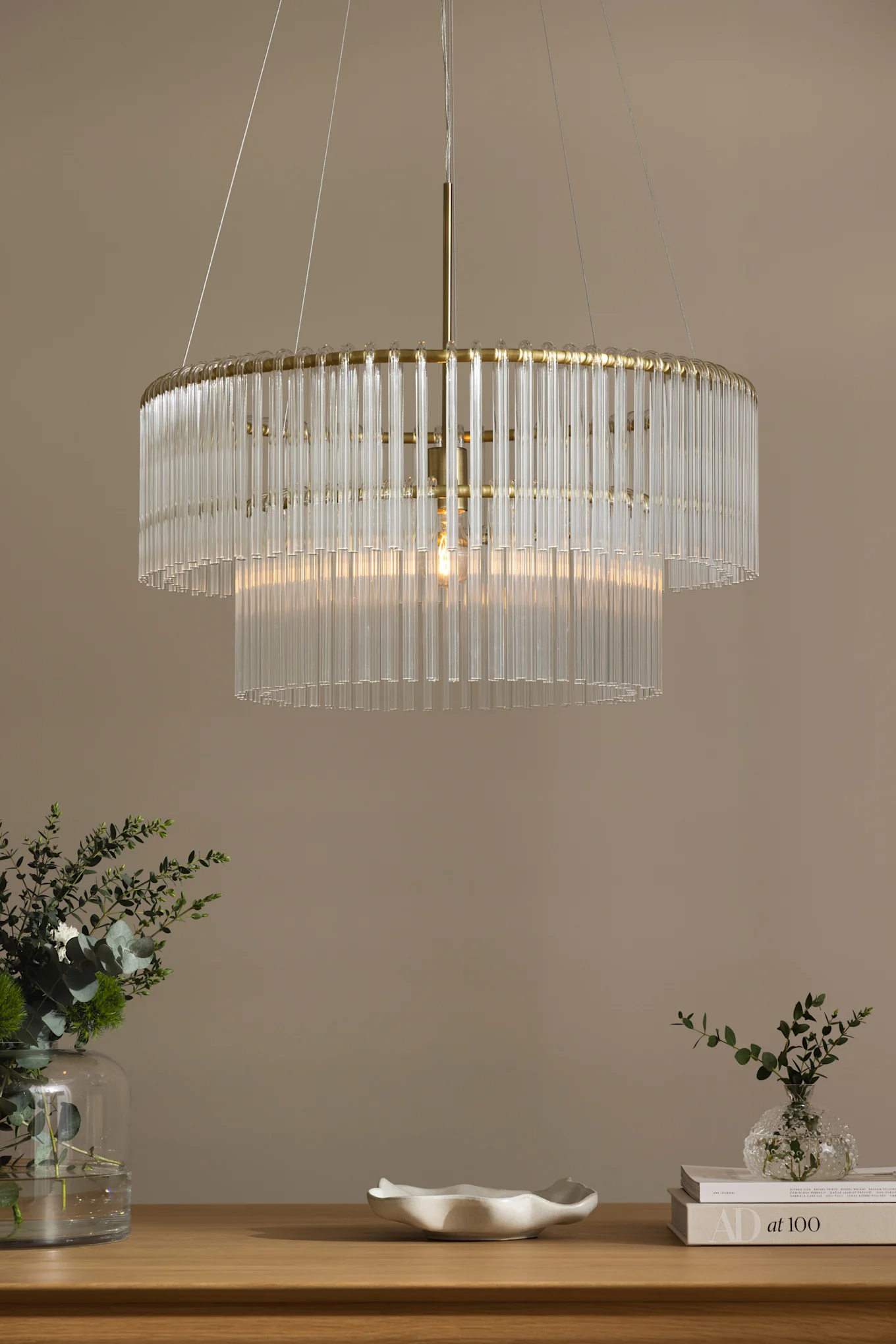 Lámpara de araña de dos niveles con varillas de cristal transparente y marco dorado, iluminando una mesa de madera con plantas verdes y un cuenco blanco.