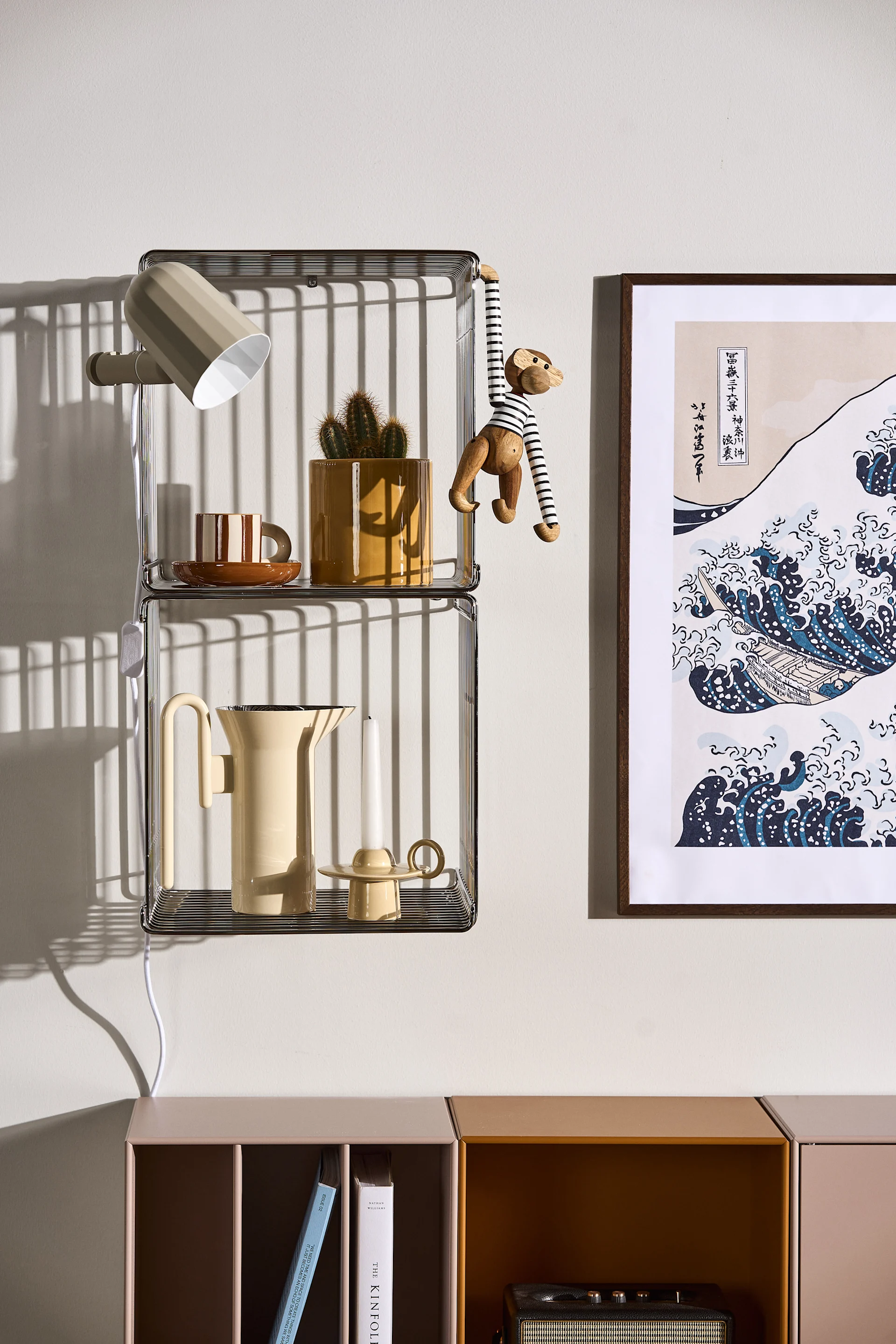 Dos estantes de rejilla metálica apilados con lámpara de pared, taza a rayas, cactus en maceta, mono de juguete, jarra y vela, junto a un grabado japonés de olas.