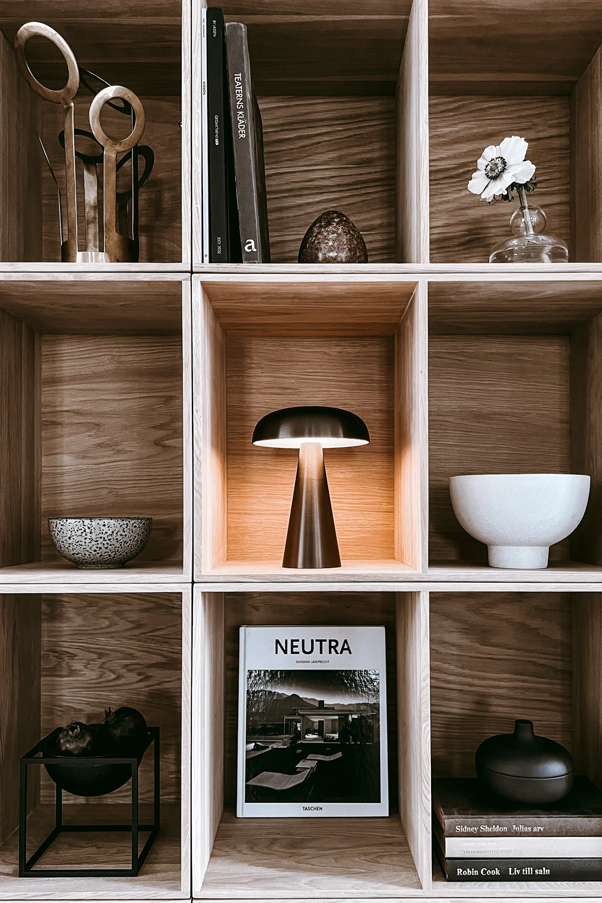 Estantería de madera con objetos decorativos: una lámpara encendida, libros, esculturas abstractas, cuencos y una flor blanca.