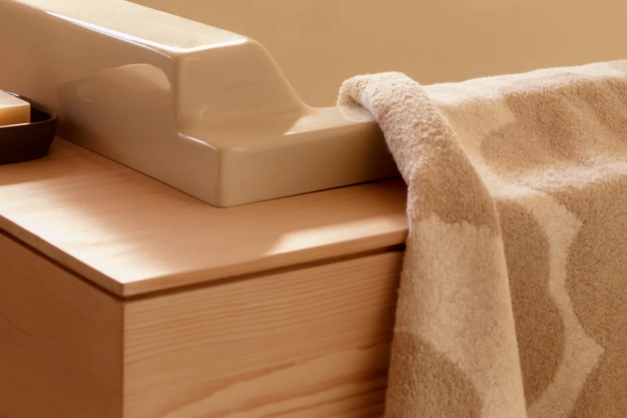 Primer plano de una toalla beige texturizada sobre un accesorio cerámico claro en un tocador de madera con una barra de jabón.