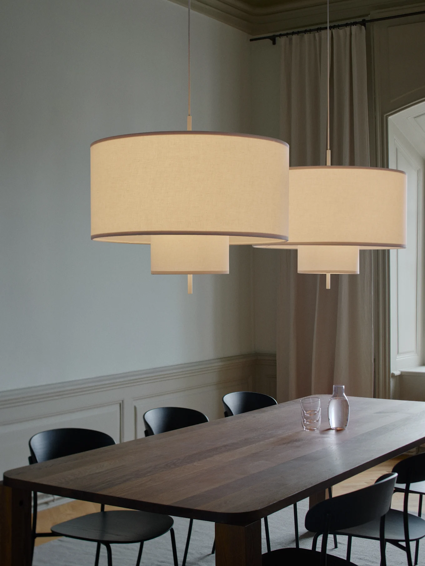 Dos lámparas colgantes de tela escalonadas iluminan una larga mesa de comedor de madera con sillas negras, una jarra y vasos.