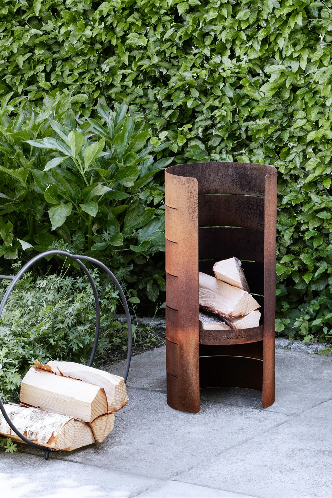 Un leñero metálico oxidado para exterior, lleno de leña, junto a un soporte circular para leña en un patio, con un seto verde al fondo.