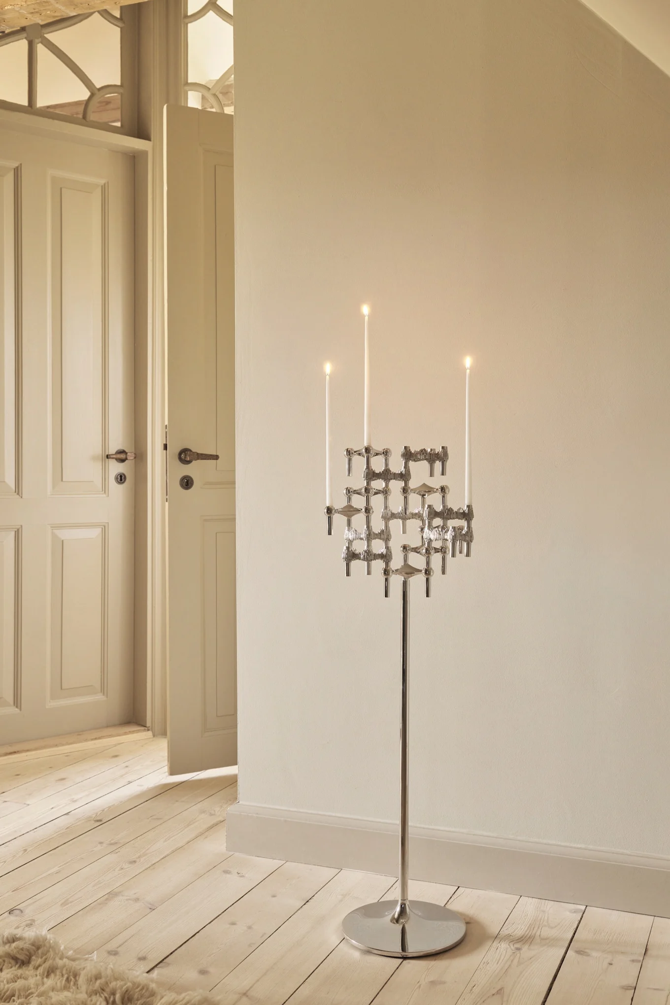 Candelabro alto de plata moderno con velas blancas encendidas sobre suelo de madera en una habitación con paredes claras y puertas abiertas.