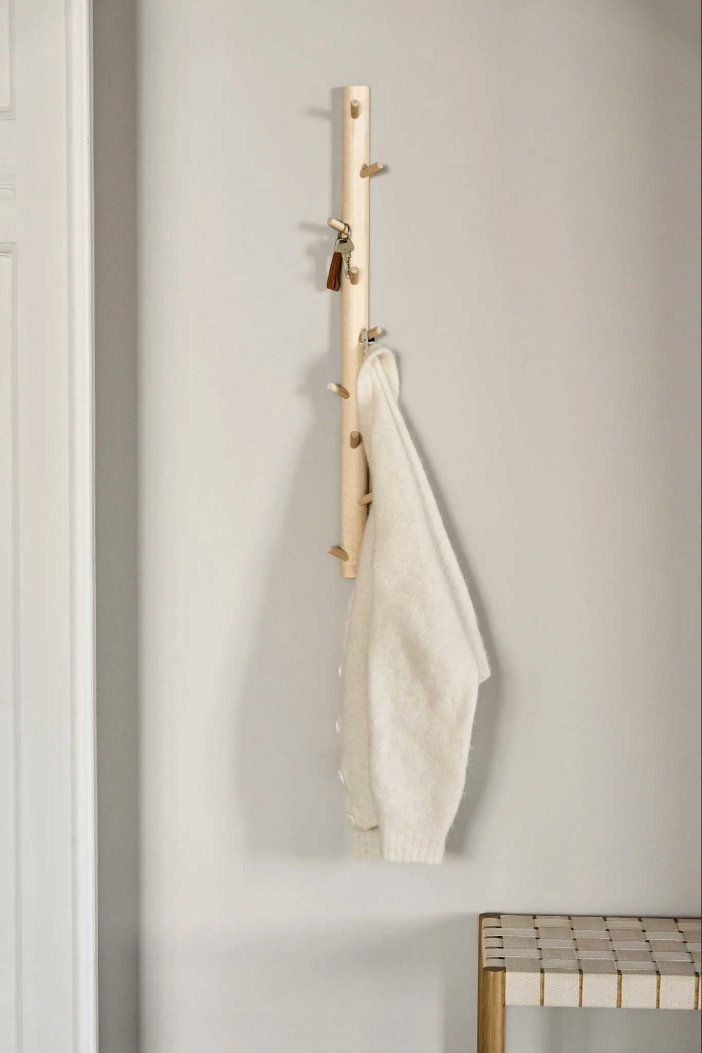 Una barra de madera clara con ganchos sostiene las llaves y un jersey blanco roto, con un banco trenzado debajo.