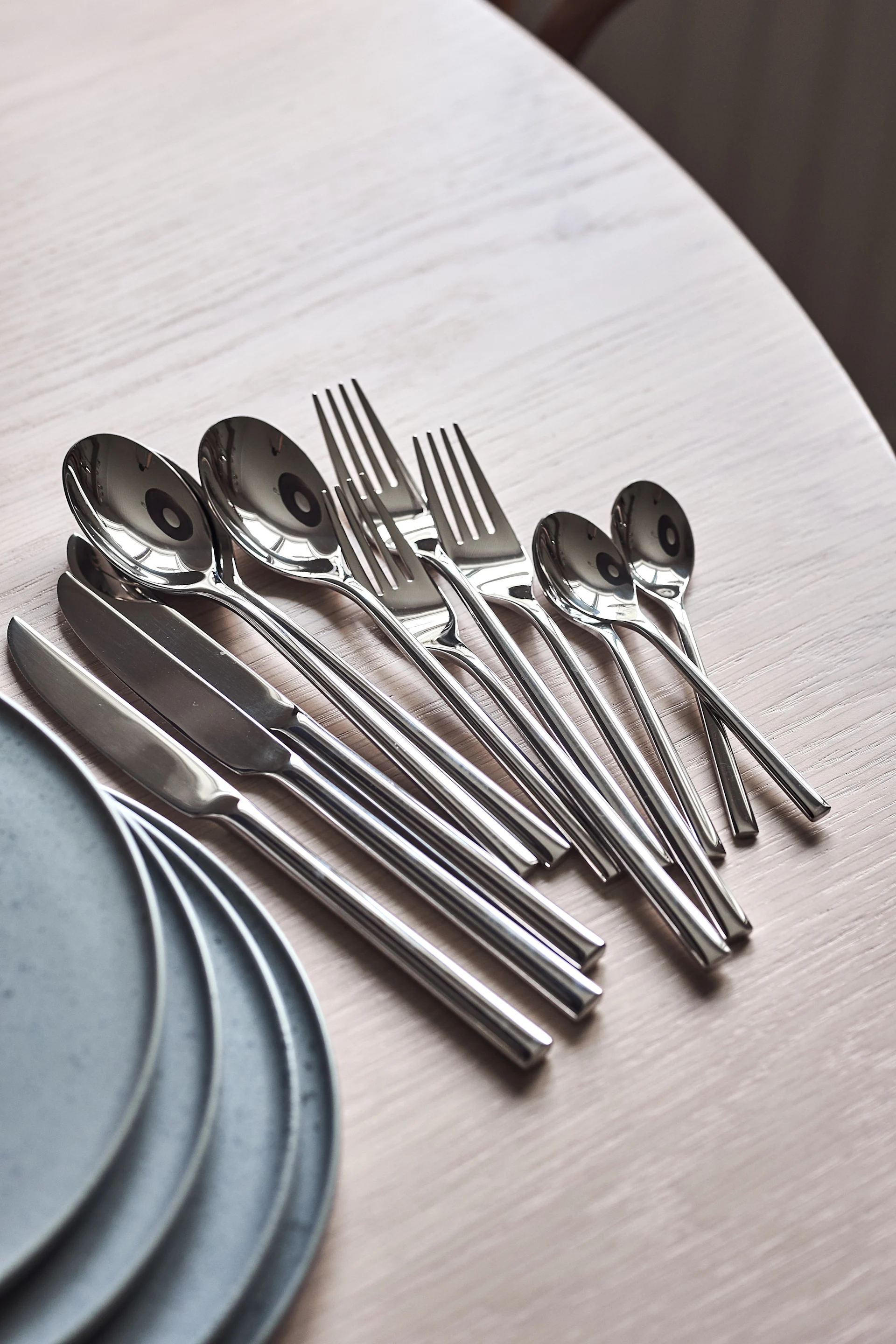 Juego de cubiertos brillantes de color plata, incluyendo cucharas, tenedores y cuchillos, dispuestos sobre una mesa de madera clara con platos grises.