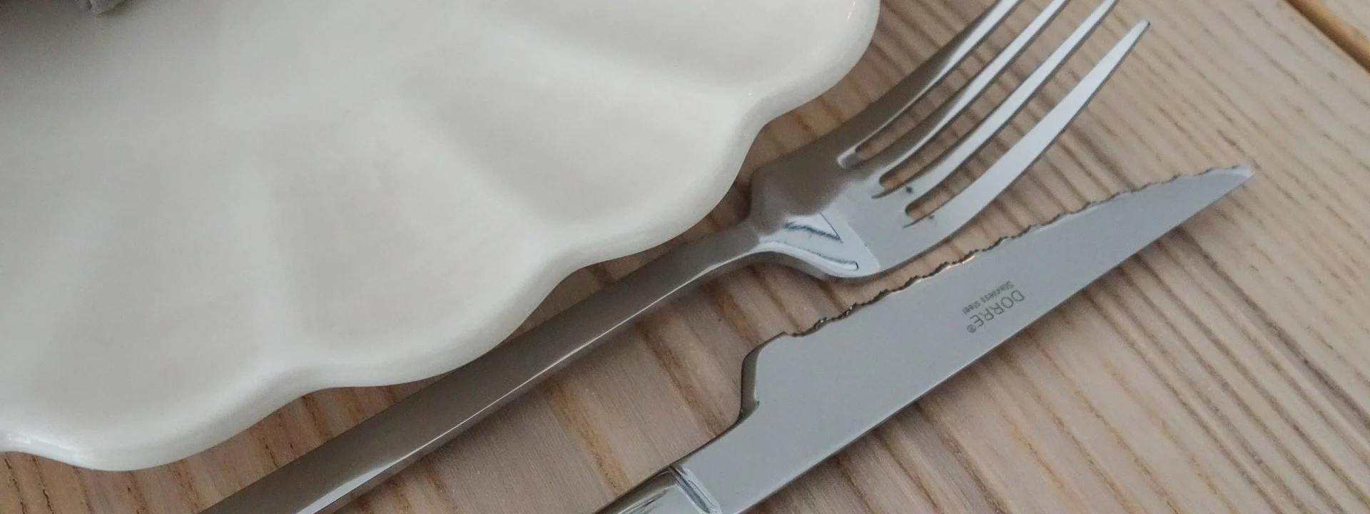 Elegante mesa puesta con plato blanco ondulado, servilleta lino gris, servilletero dorado, tenedor y cuchillo plateados, copa vino en mesa madera.