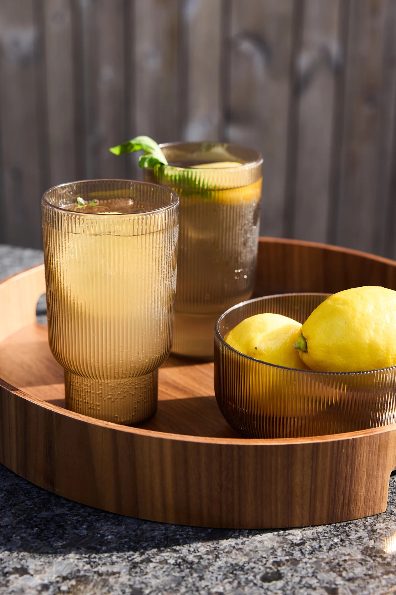 Dos vasos acanalados ámbar con limonada y un cuenco de limones en una bandeja de madera al aire libre.
