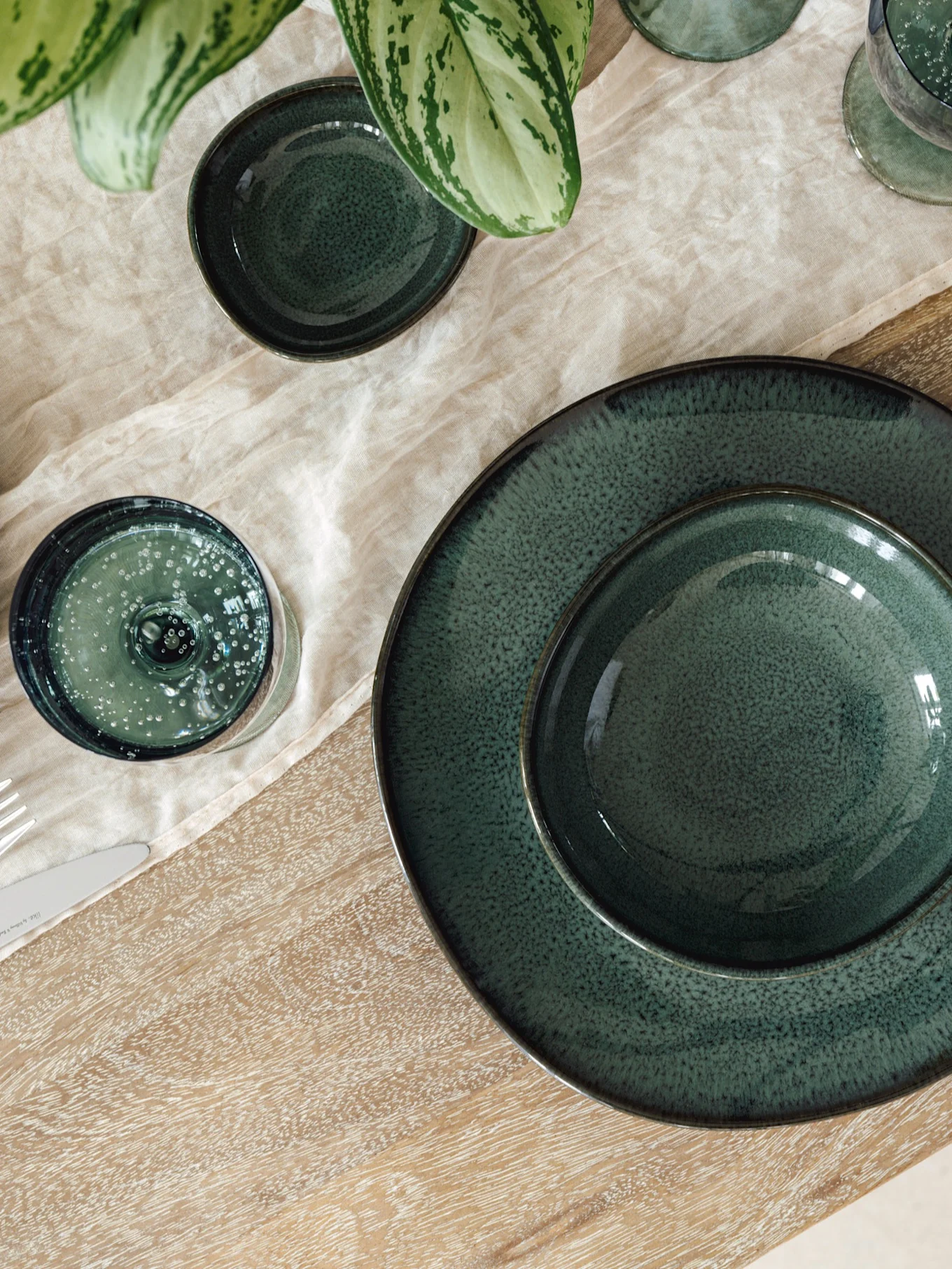 Vajilla verde oscuro de cerámica con platos, cuencos y vasos sobre mantel de lino claro y mesa de madera, con planta.