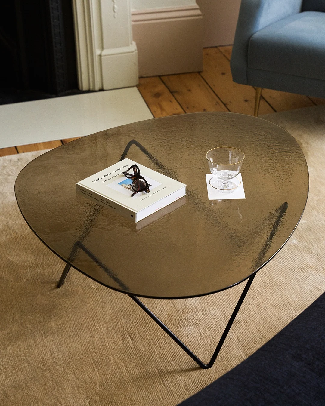 Mesa de centro de cristal oscuro con forma orgánica, con un libro, gafas de carey y un vaso con borde dorado sobre un posavasos.