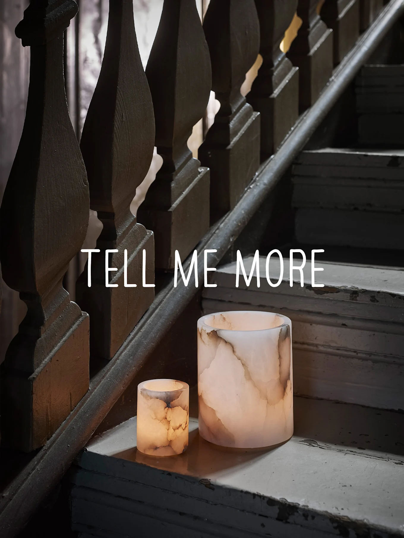 Dos candelabros luminosos tipo ónix en una escalera de madera gastada, con el texto "TELL ME MORE".