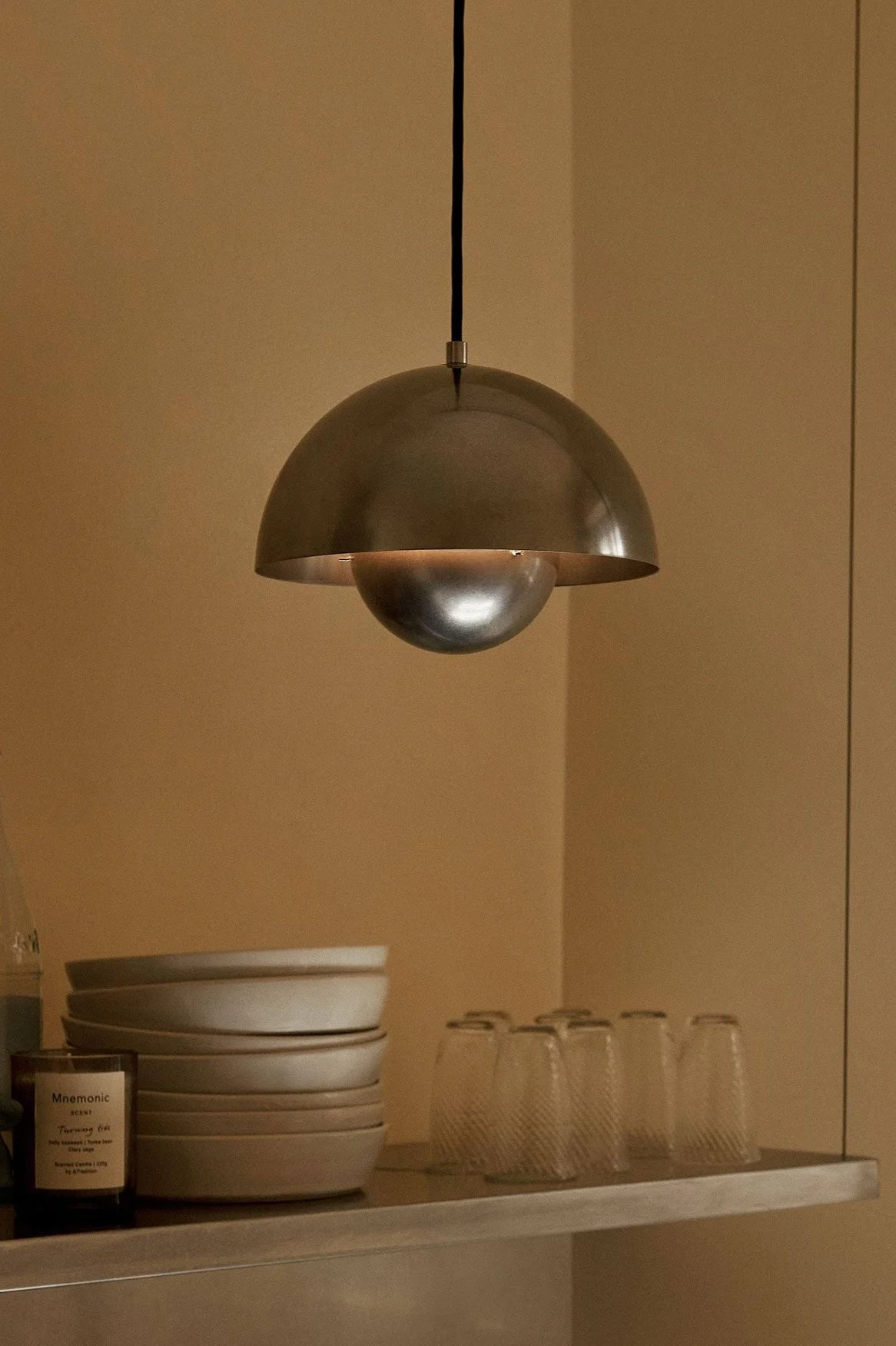 Una lámpara colgante de metal en forma de cúpula cuelga sobre un estante con cuencos, vasos, botellas y una vela sobre una pared marrón cálida.