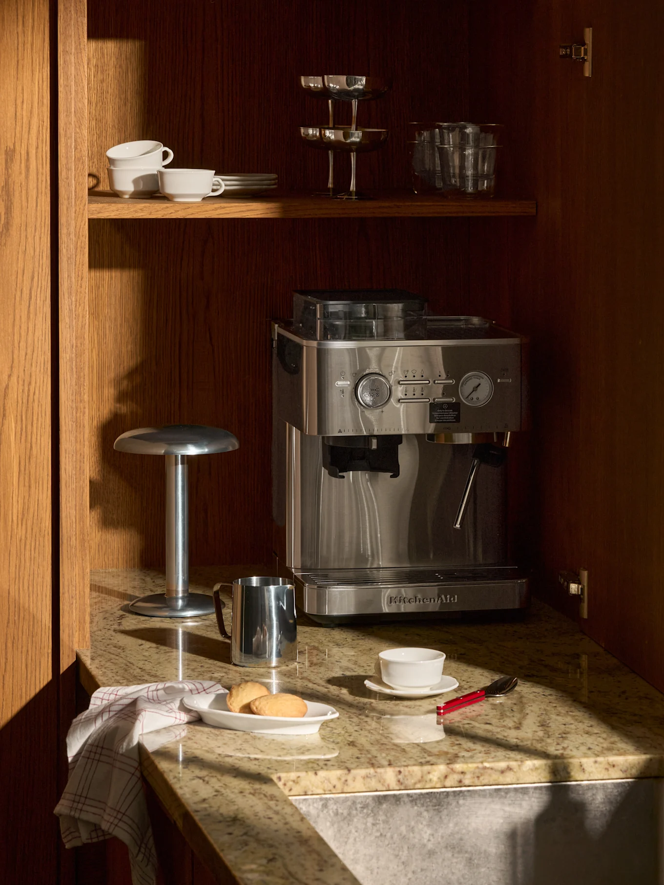 Cafetera espresso de acero inoxidable en encimera de granito, armario de madera. Arriba tazas y vasos. En encimera, galletas, taza, jarra de leche.