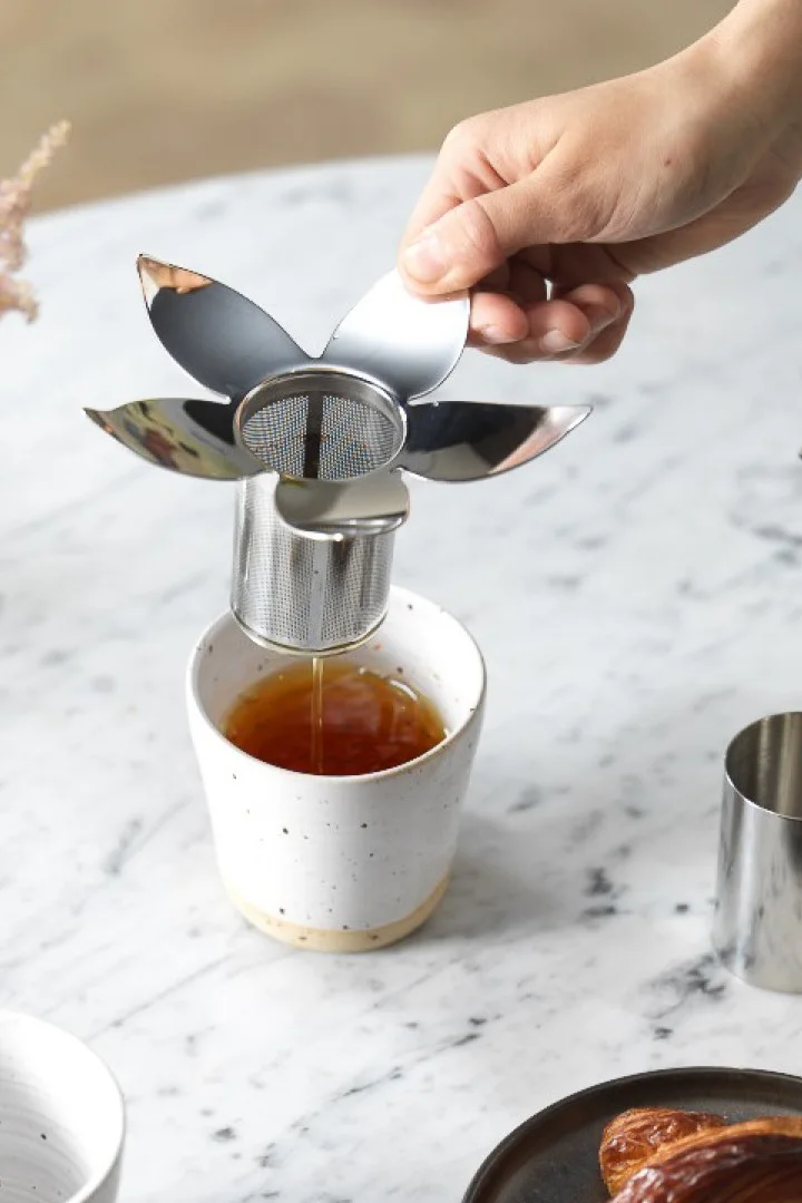 Mano sosteniendo un infusor de té en forma de flor, goteando té en una taza de cerámica blanca sobre una superficie de mármol.