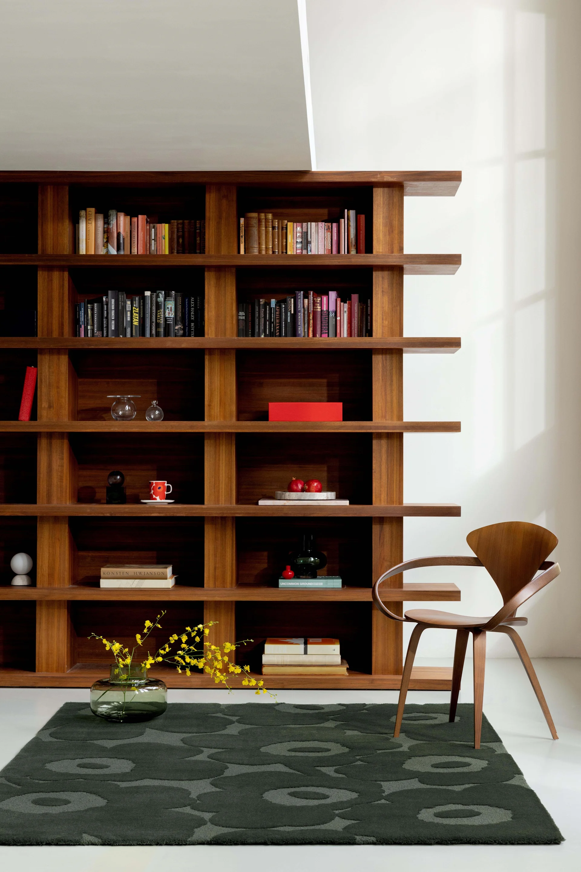 Una habitación elegante con una alta estantería de madera llena de libros y decoración, una silla de madera curva y una alfombra verde oscuro.