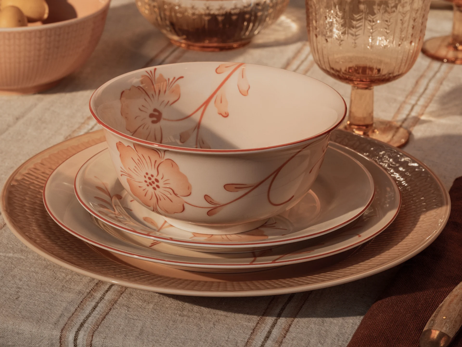 Primer plano de un ambiente de mesa rústico con platos apilados de color melocotón y crema, una copa ámbar y un queso blanco acanalado sobre un mantel de lino a rayas.