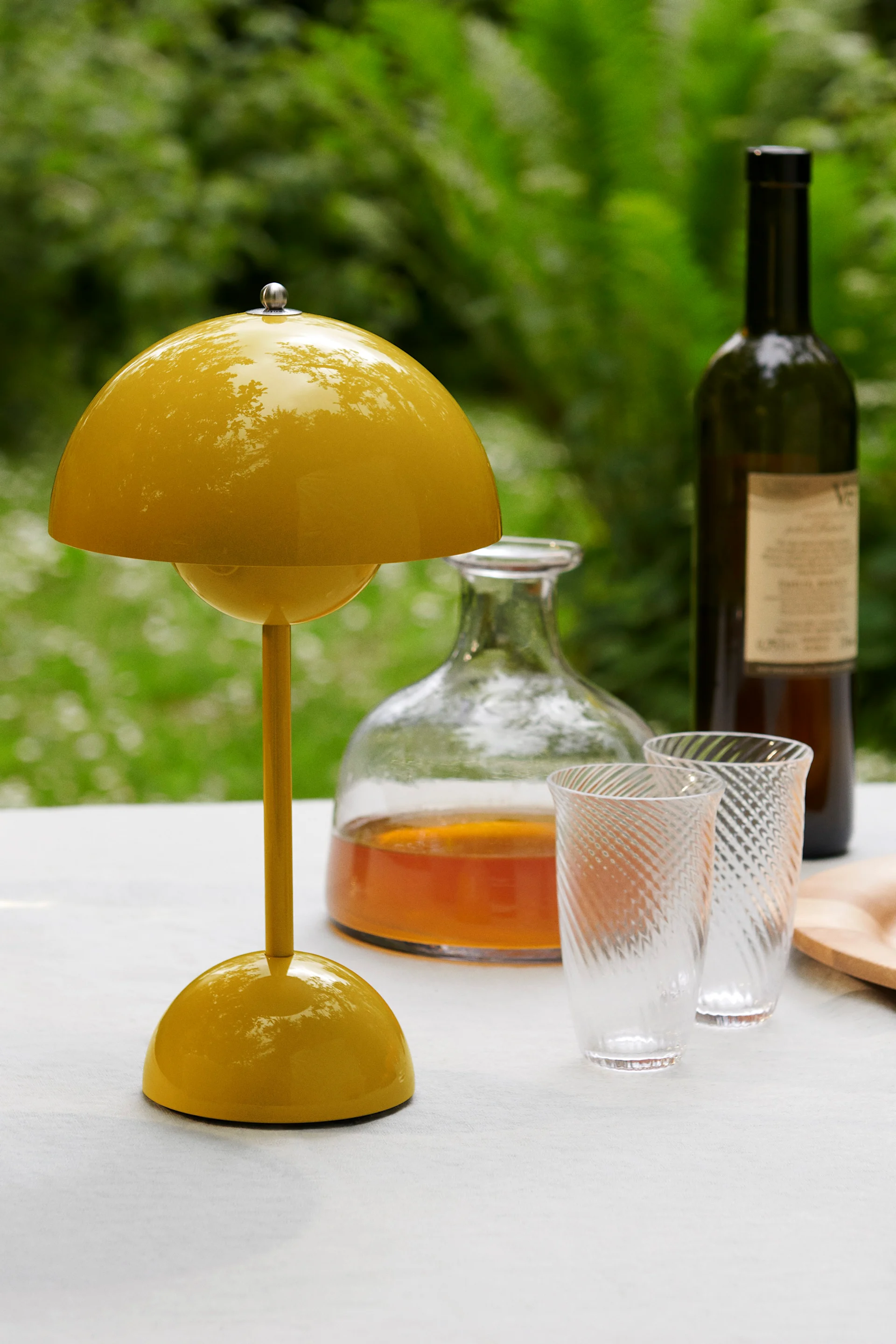 Una lámpara de mesa amarilla brillante en forma de seta sobre una mesa blanca, con una jarra, dos vasos estampados y una botella de vino detrás, sobre un fondo verde.