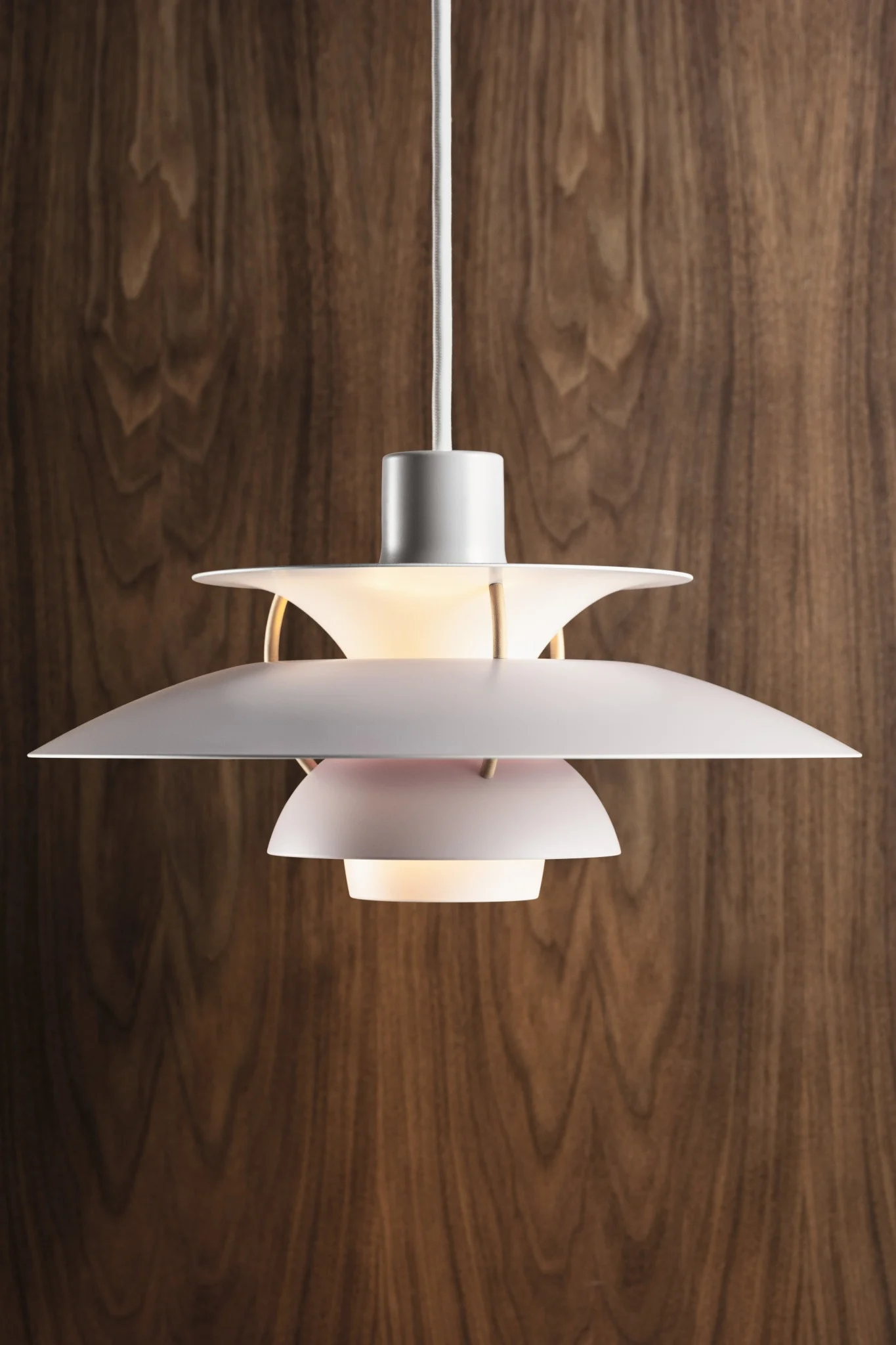 Una lámpara colgante blanca de varios niveles con un brillo cálido, colgada sobre una pared de paneles de madera oscura.