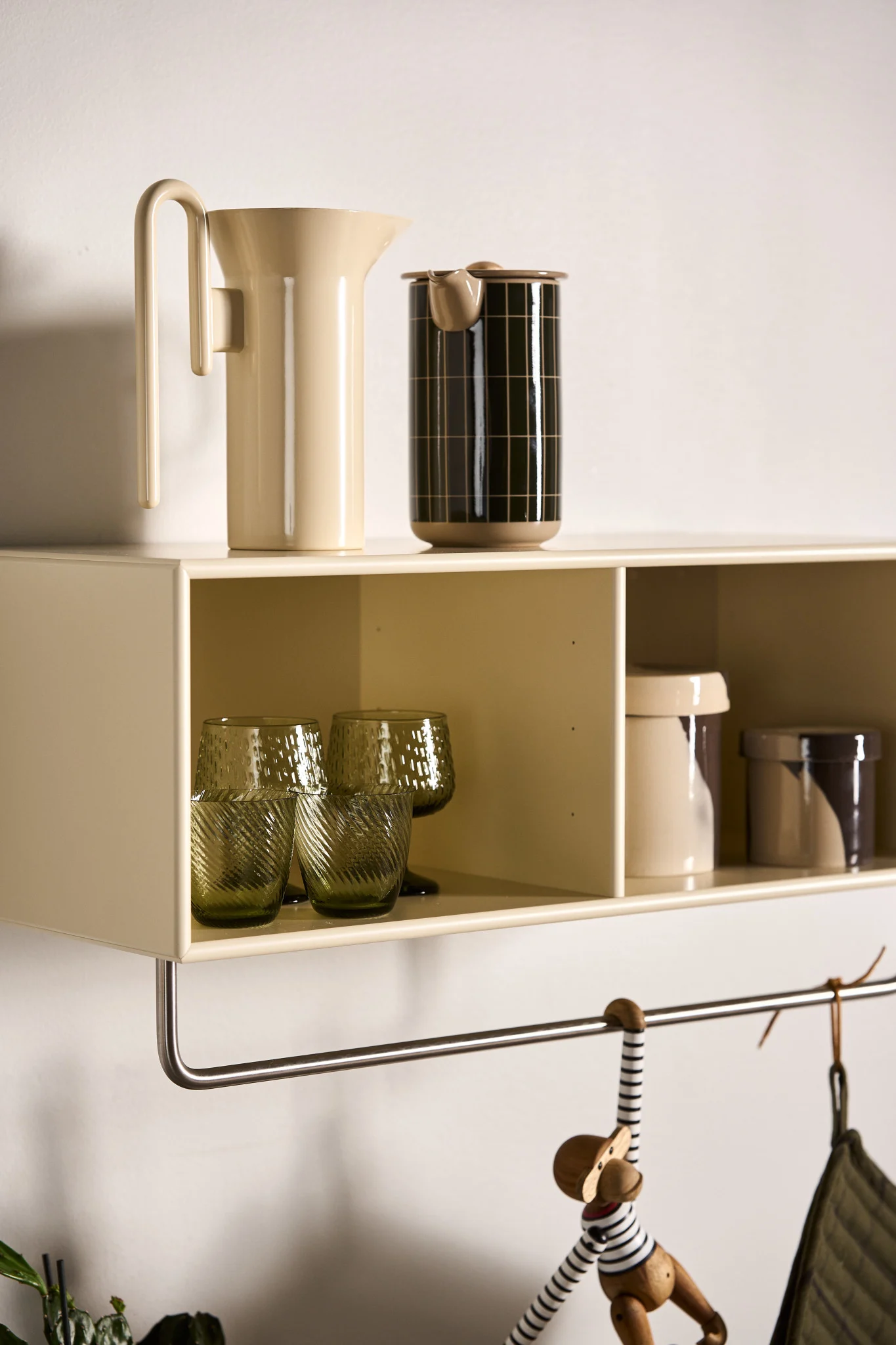 Un estante de pared beige claro con una jarra crema, un recipiente negro, vasos verdes texturizados y botes estampados. Un mono de madera cuelga debajo.