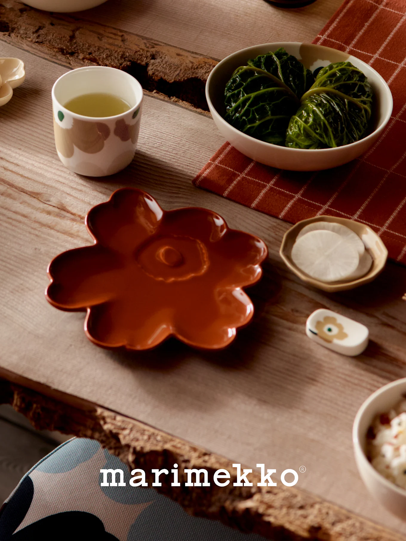 Mesa puesta con vajilla Marimekko: un plato floral color óxido, una taza de té verde, rollos de col y rábanos en rodajas.