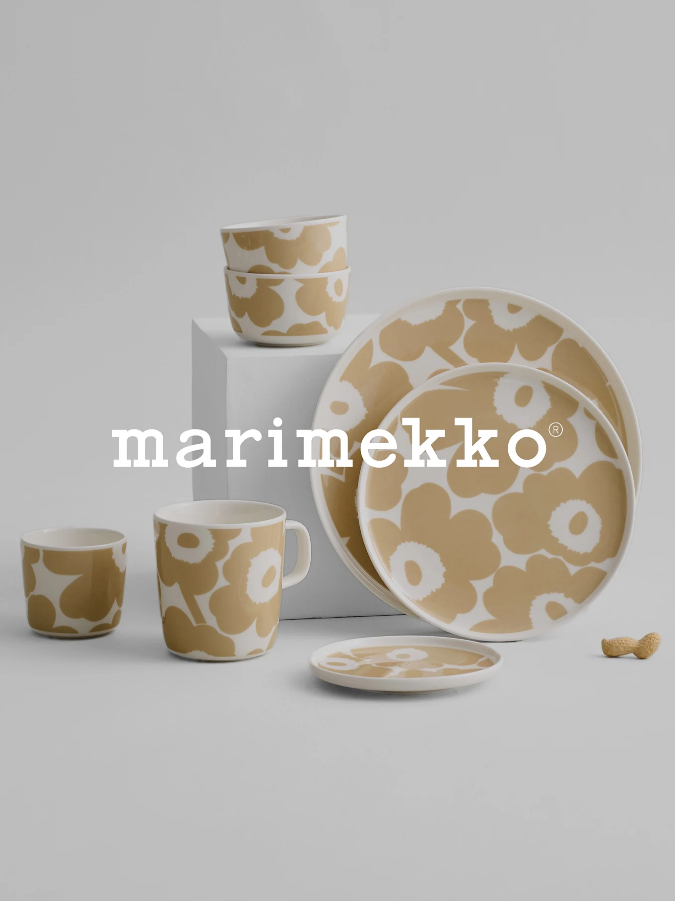 Juego de vajilla Marimekko con diseño floral beige y blanco, con cuencos, taza, vasos, platos y un cacahuete sobre una superficie gris.