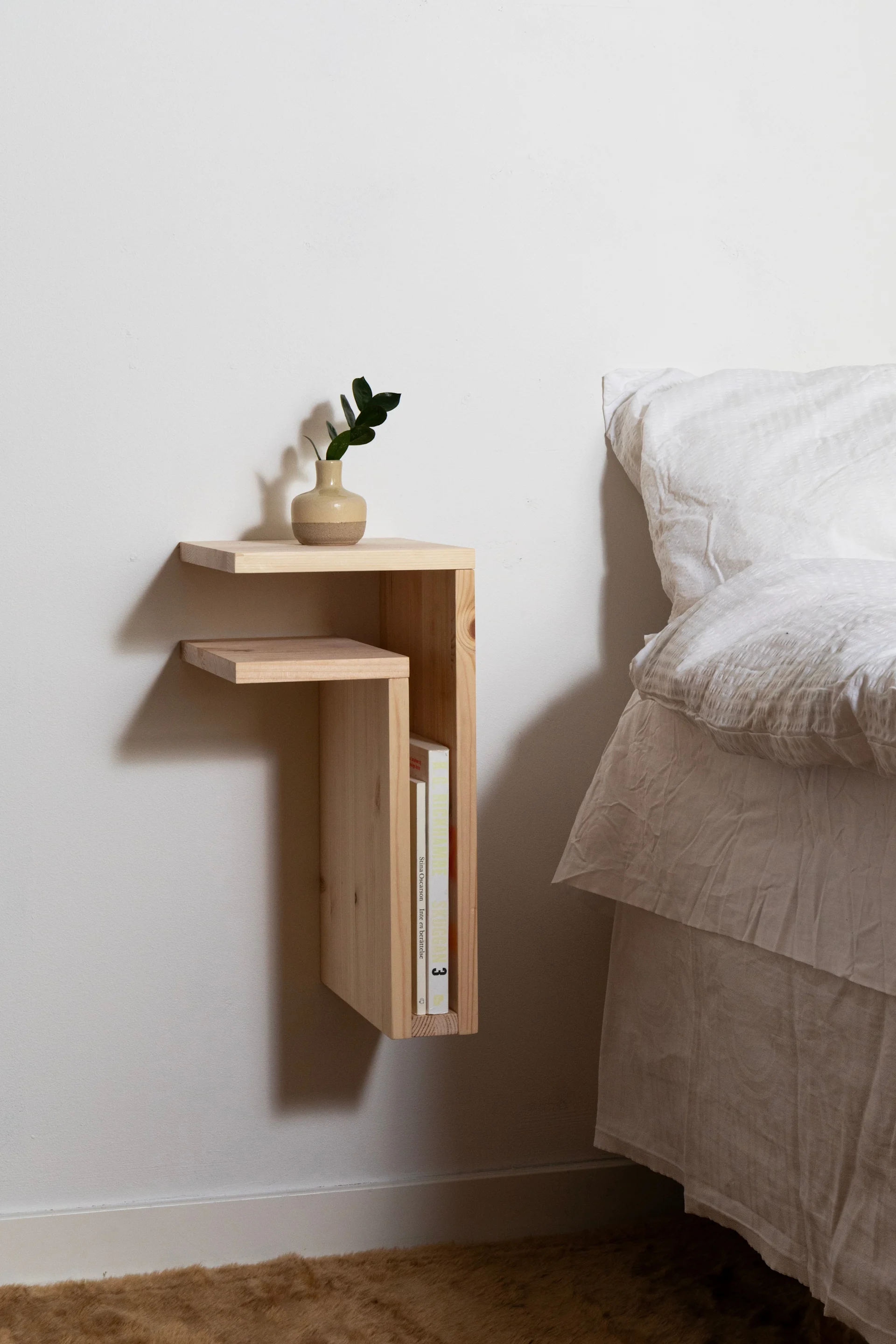 Una mesita de noche flotante de madera clara con una planta en un jarrón de dos tonos y libros, junto a una cama con ropa de cama blanca.