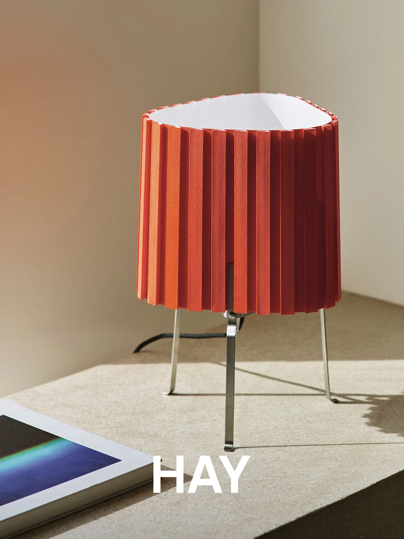 Lámpara de mesa naranja plisada con base de metal sobre una superficie texturizada, junto a un libro. La palabra 'HAY' está superpuesta.