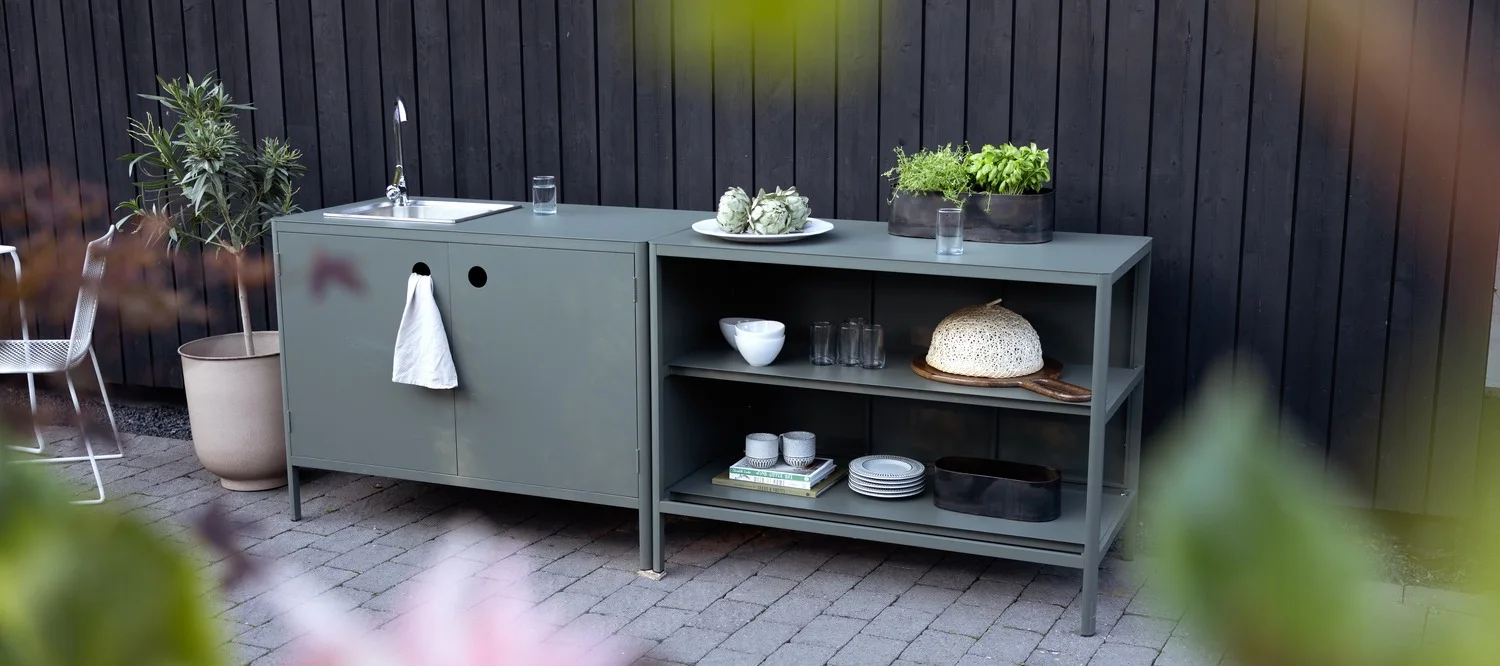 Una  cocina exterior en color verde grisáceo, con fregadero y toalla a la izquierda, y estantes con menaje y hierbas en maceta a la derecha.