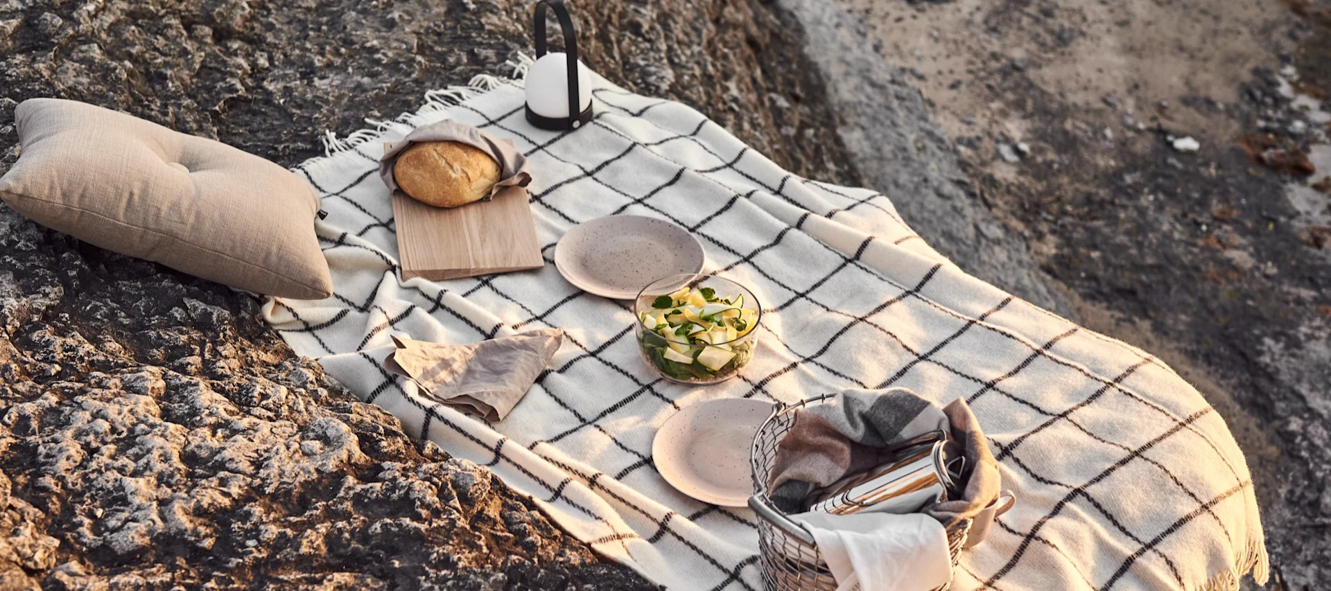 Un set de picnic sobre rocas, con una manta de cuadros blanca, un cojín, pan, un tazón de ensalada, platos, un farolillo y una cesta con una manta.