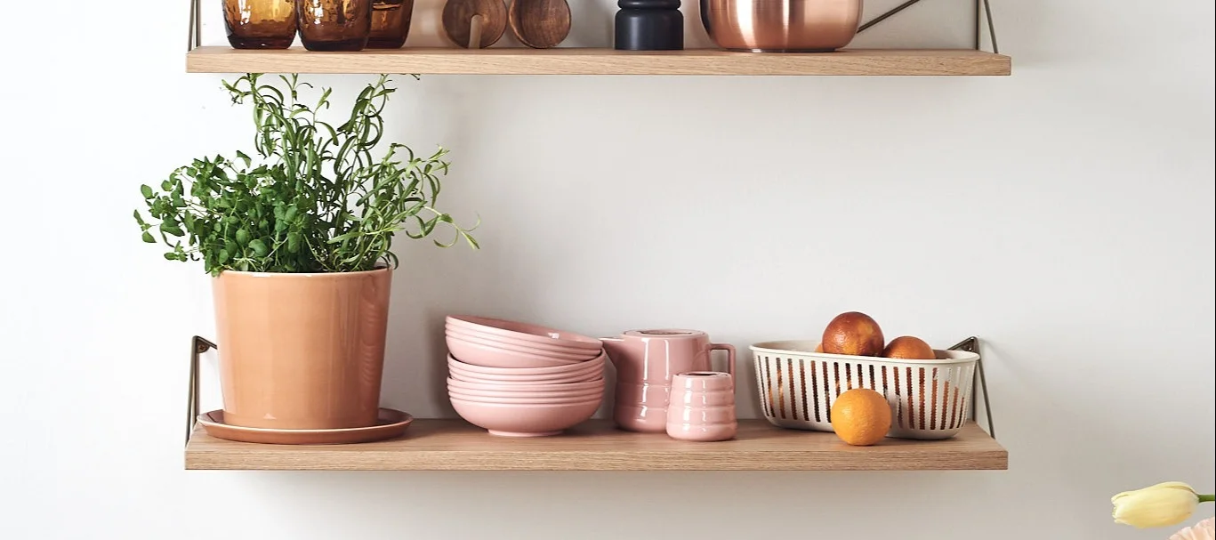 Dos estanterías de color burdeos junto a una pared clara, con libros, esculturas de color beige y una lámpara de color naranja brillante con pantalla roja.