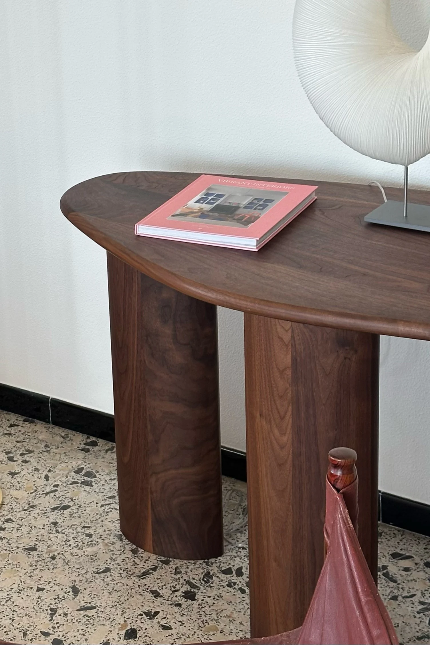 Mesa auxiliar moderna de madera oscura con un libro rosa y una lámpara blanca plisada sobre un suelo de terrazo.