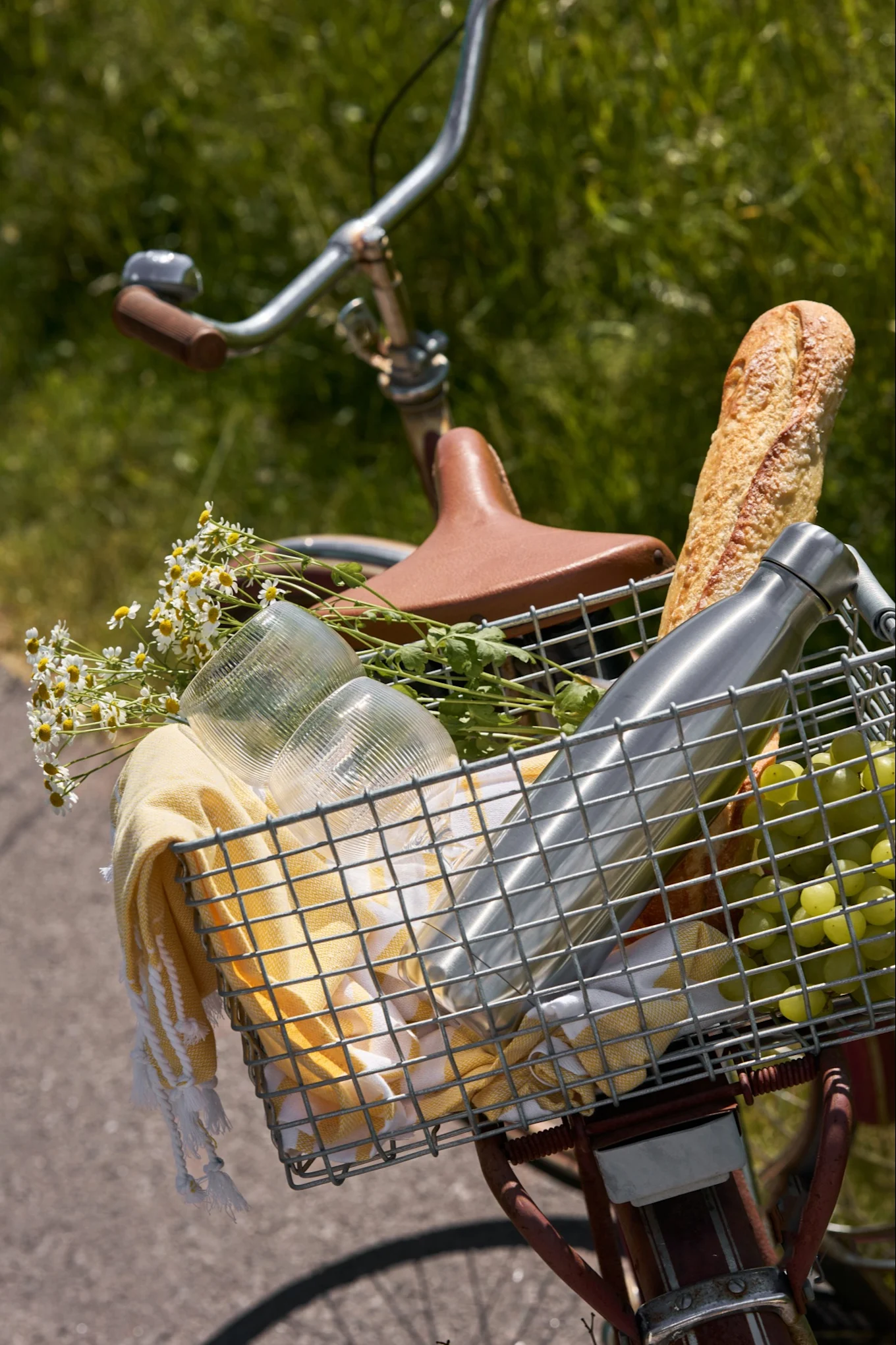 Cesta delantera de bicicleta con artículos de picnic: una baguette, botella de agua, uvas, vasos, toalla amarilla y margaritas blancas.