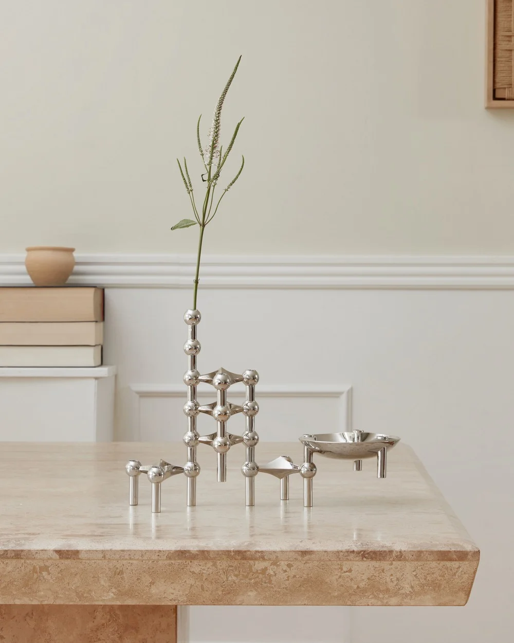 Un sistema modular de candelabros de plata pulida con un tallo de planta y un pequeño plato sobre una mesa de travertino.