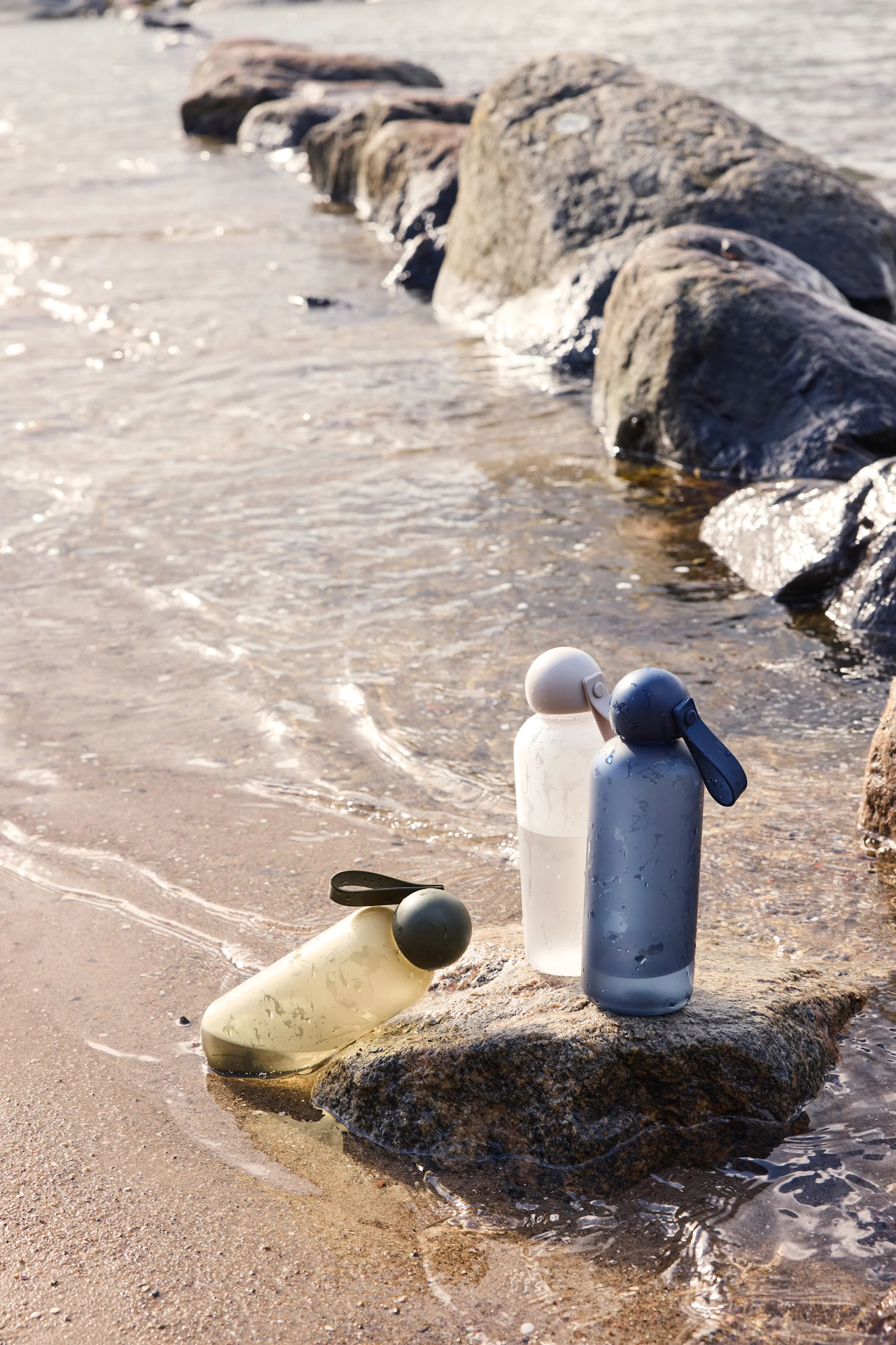 Tres botellas de agua reutilizables en amarillo, blanco esmerilado y azul grisáceo sobre una roca junto al agua.